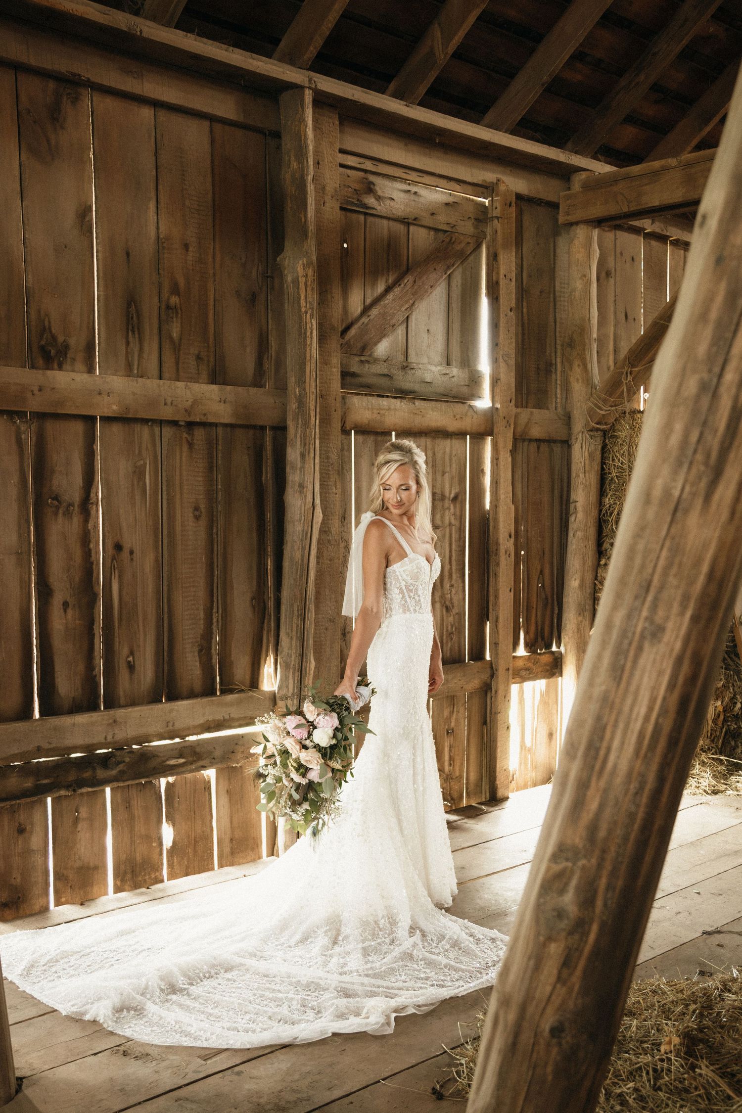Bride in lace mermaid wedding gown holding floral bouquet standing in rustic wooden barn with natural light.