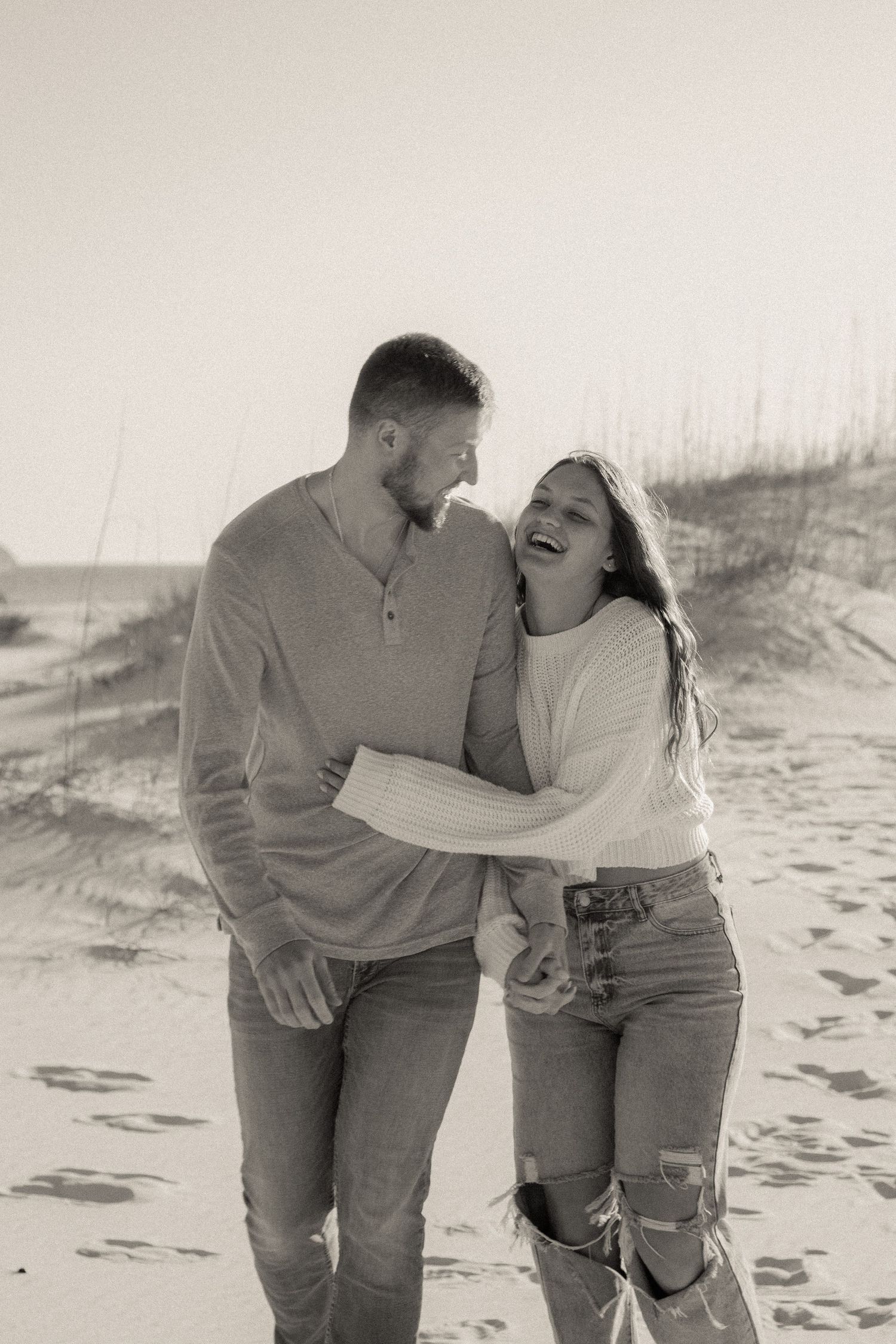 Playful couple laughing together on sandy beach during casual photoshoot.