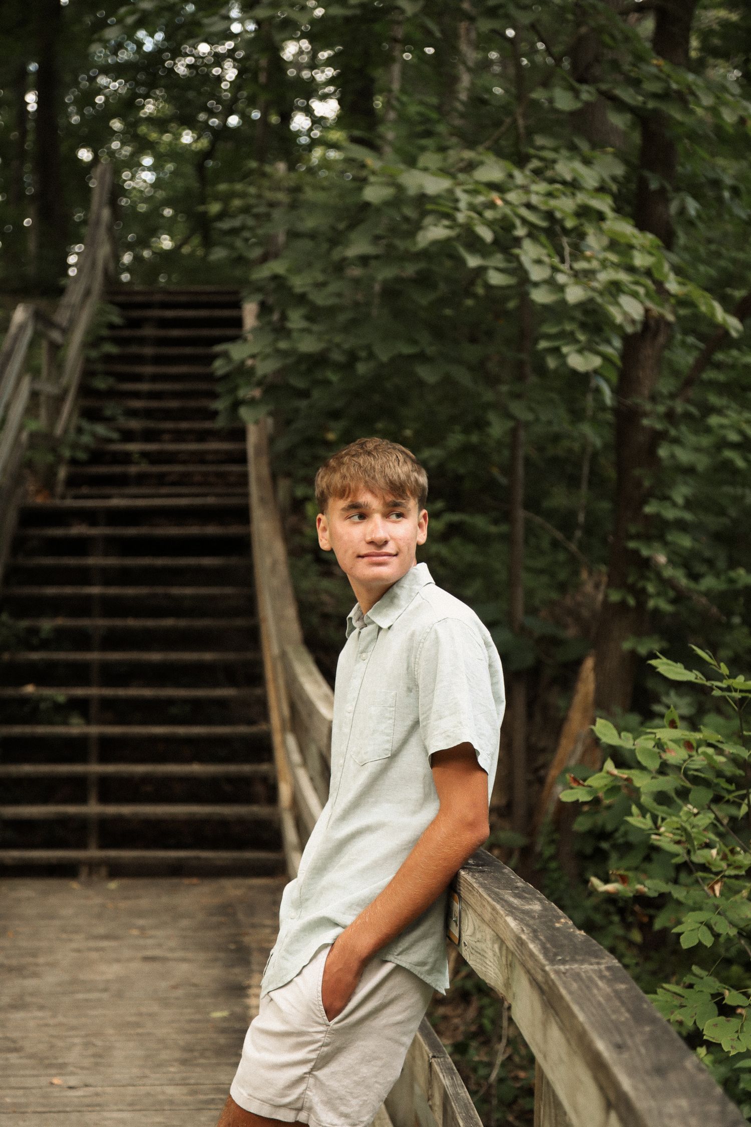 Young person in casual attire poses on wooden stairs surrounded by lush greenery.