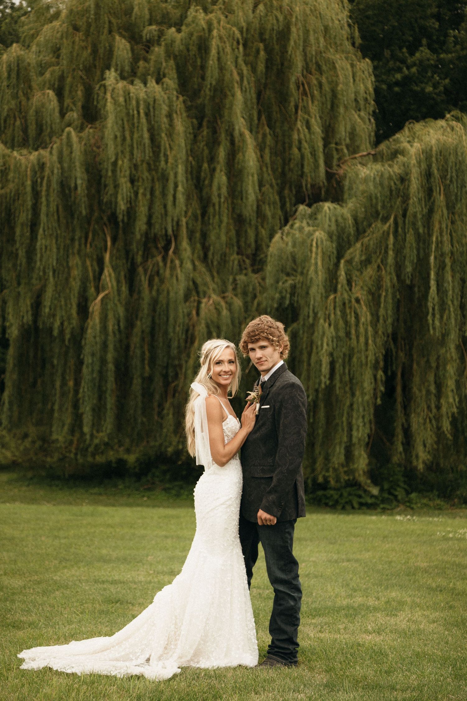 Bride and groom posing together on green lawn in front of a large weeping willow tree.