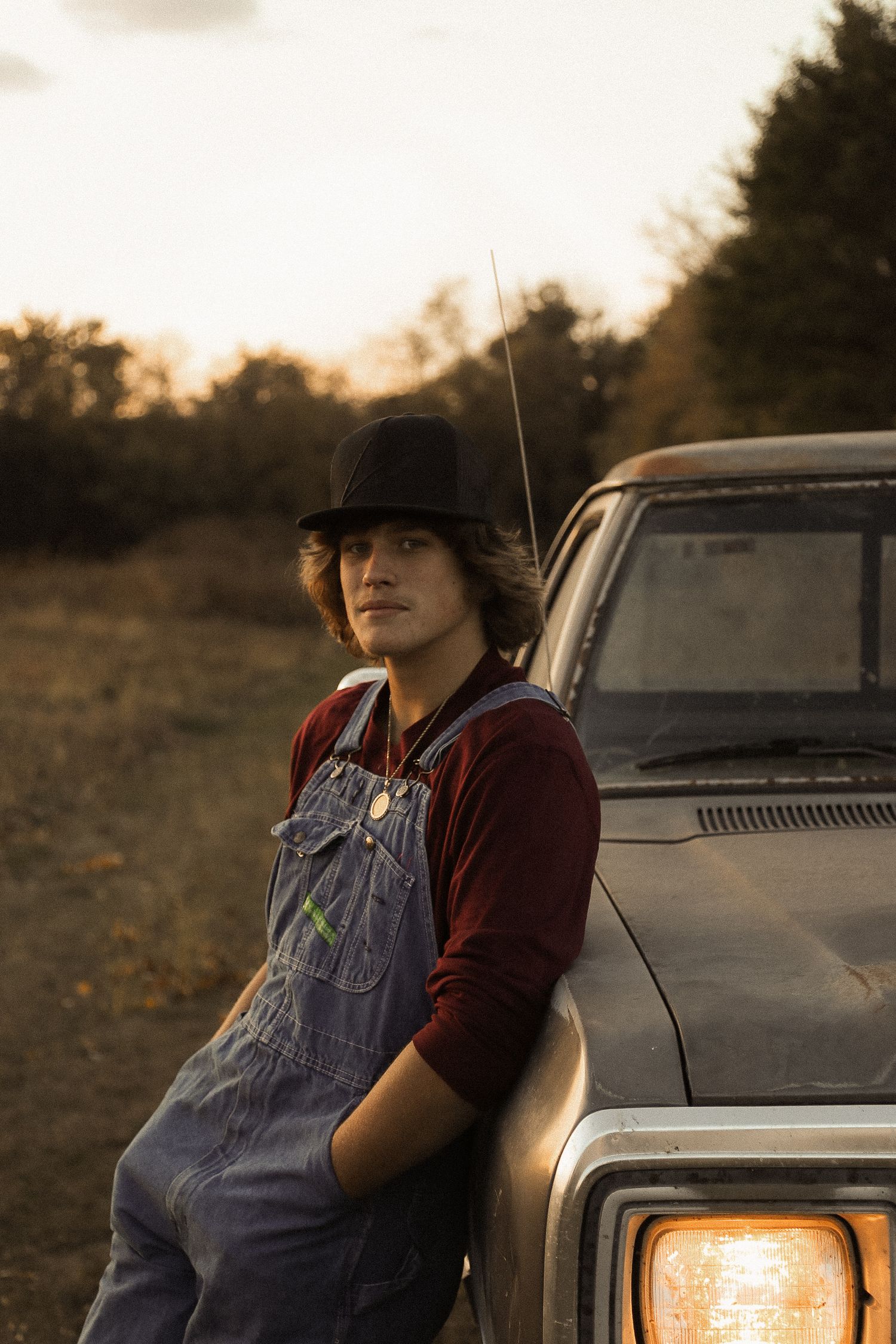 Person in overalls and burgundy shirt leans against vintage Dodge truck at sunset.