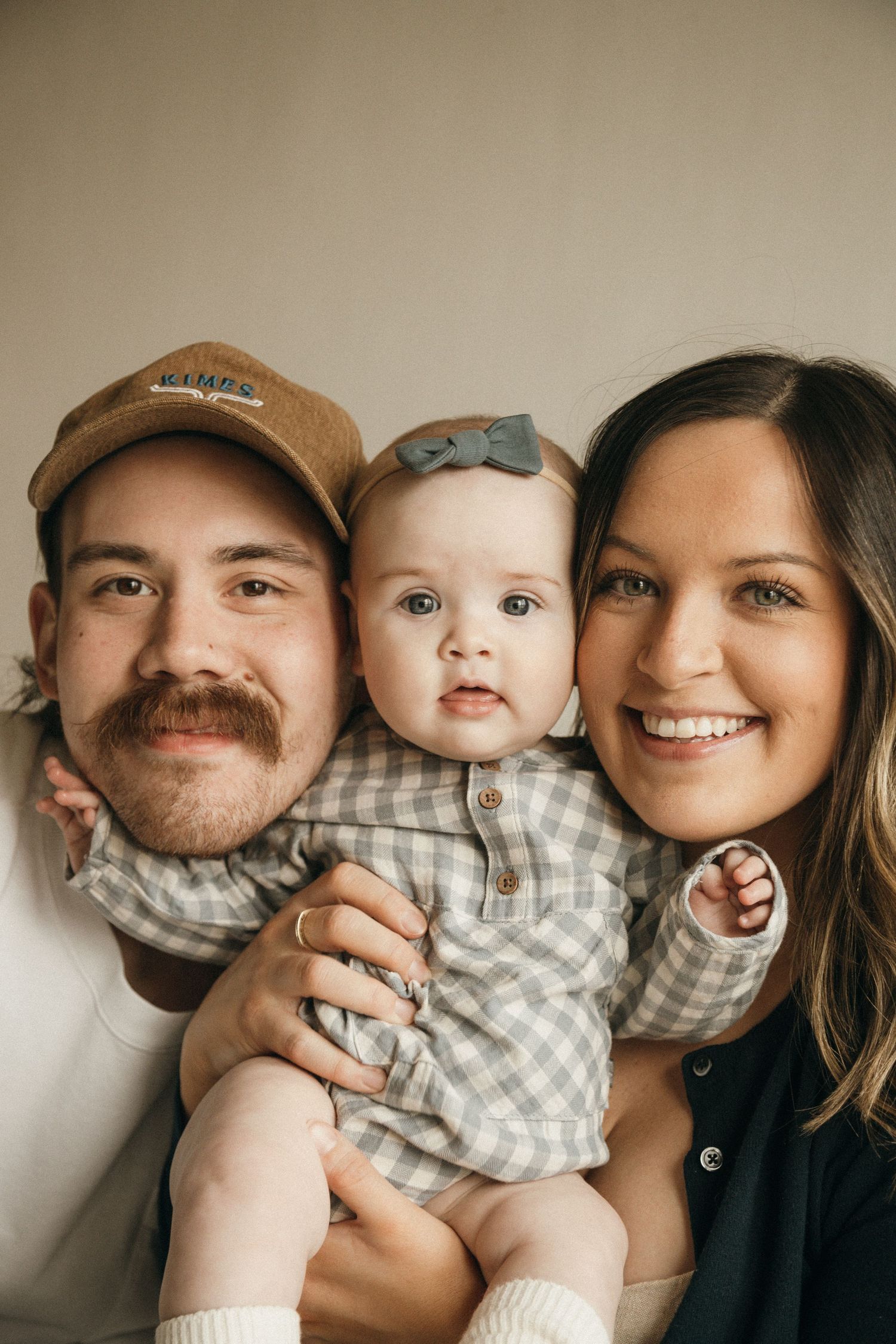 A smiling couple holds their adorable baby dressed in a gray gingham outfit with a bow headband for a family portrait.