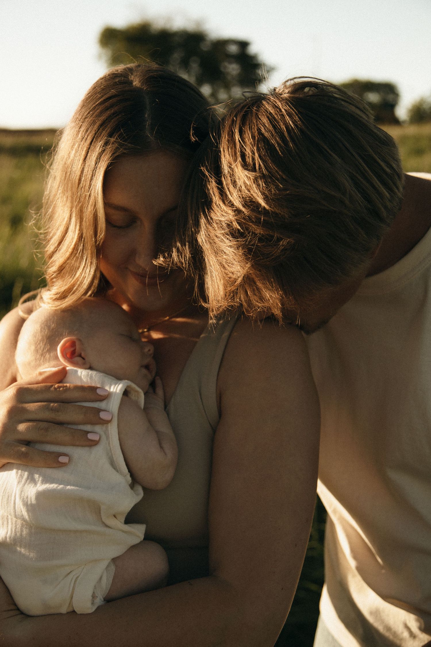 Parents sharing an intimate moment, gently holding their sleeping newborn in warm sunset light.