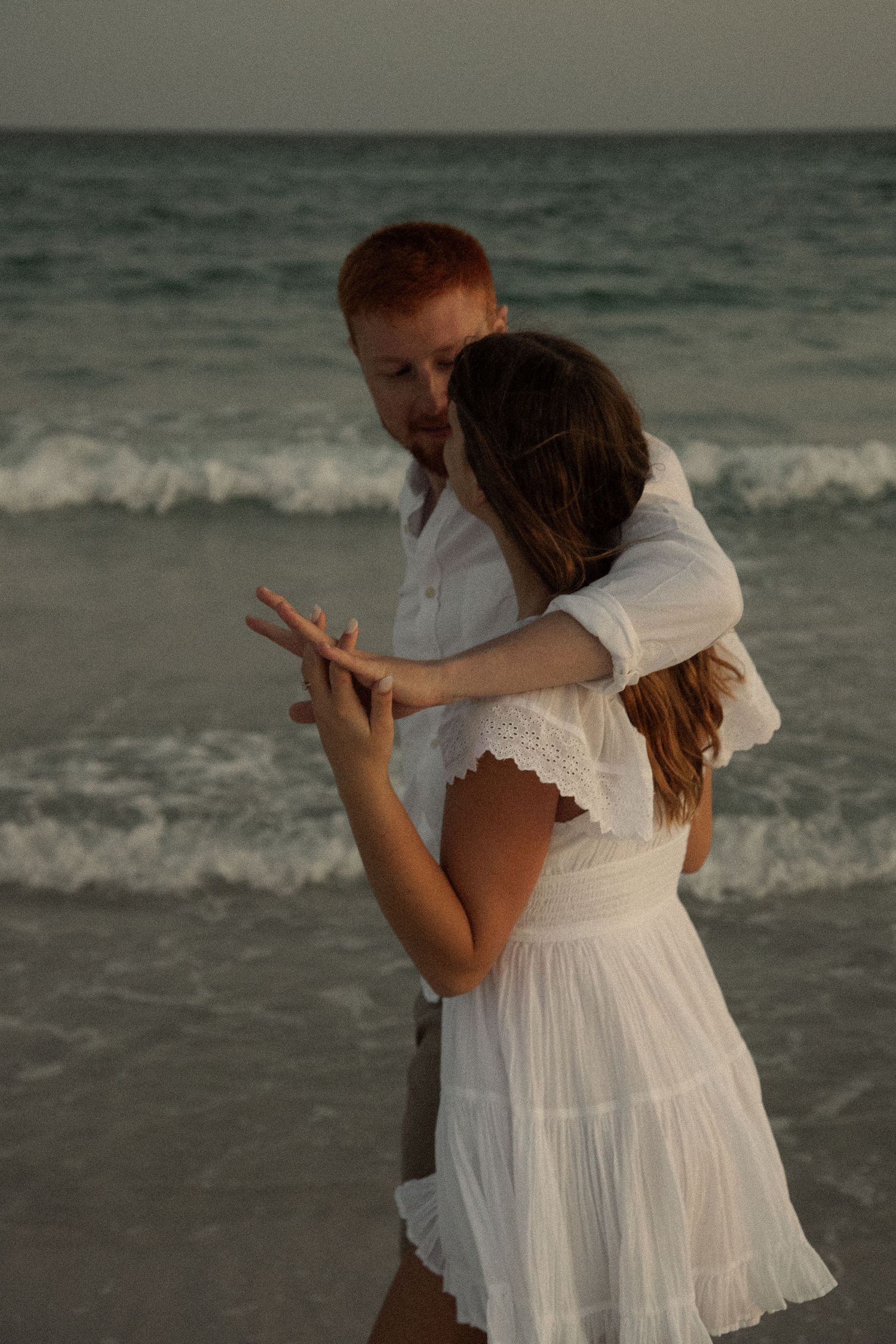 A playful couple dances on the beach at sunset with crashing waves behind them.