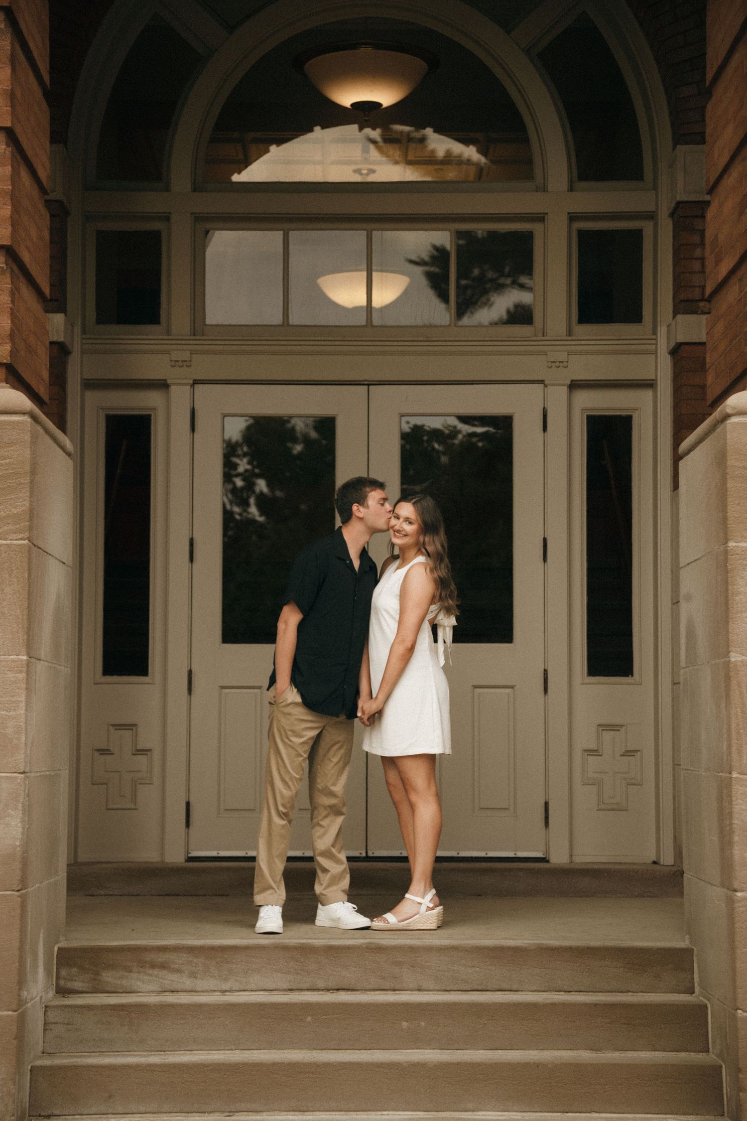 Couple holding hands and kissing on steps of elegant building with arched doorway.