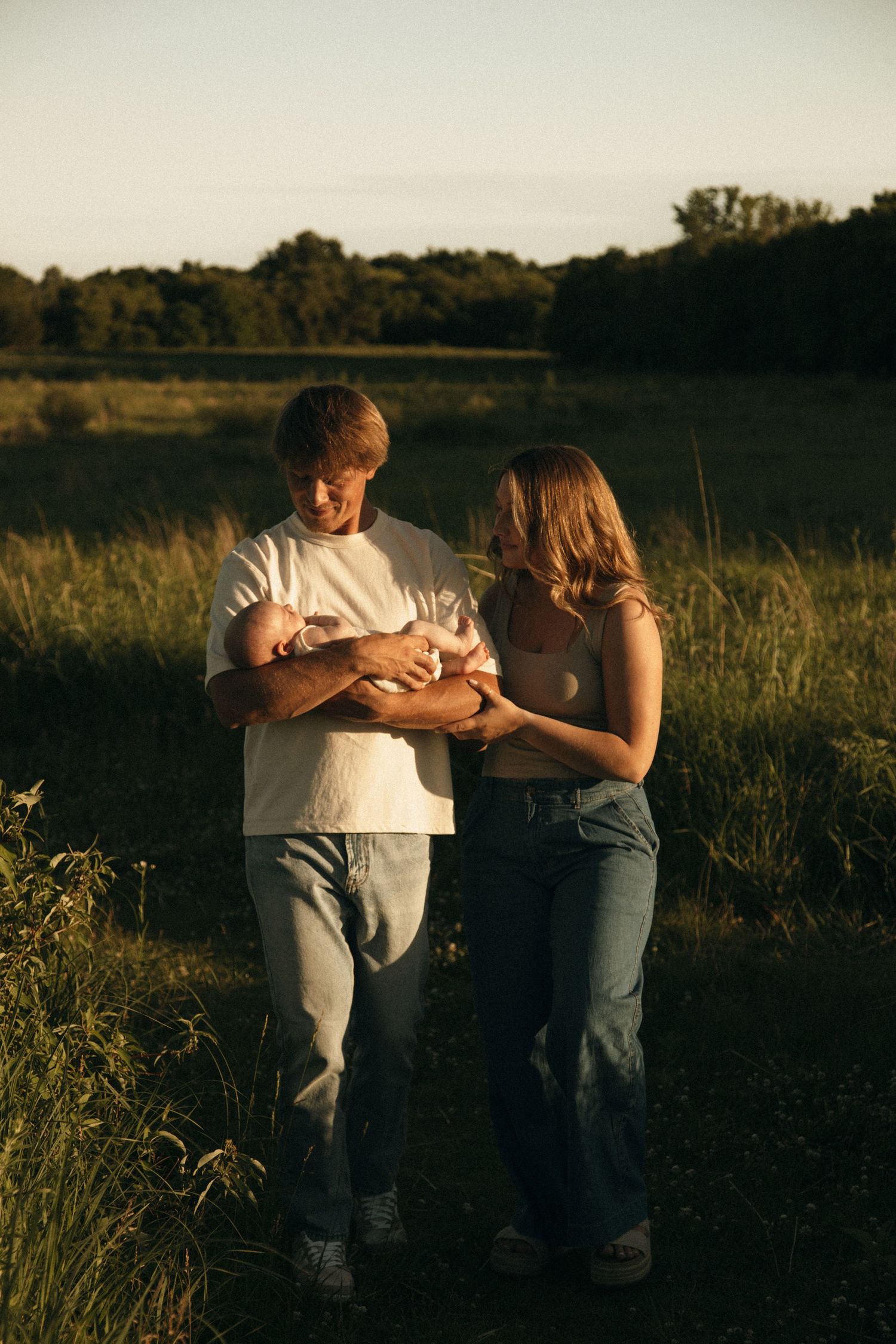New parents walking through a sunlit meadow, lovingly gazing at their newborn baby.