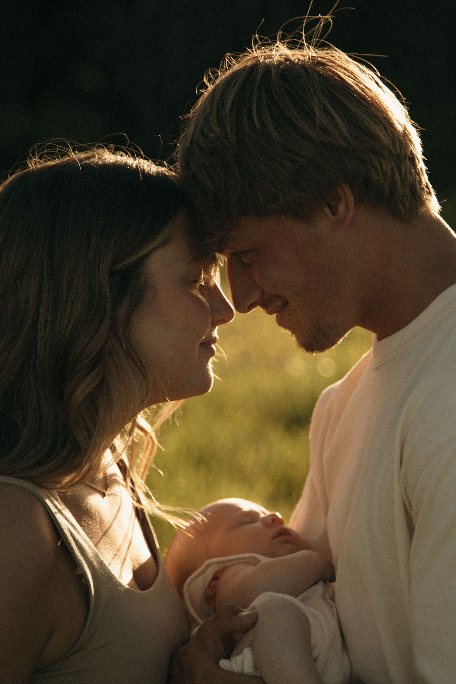 A couple tenderly touches foreheads while cradling their newborn baby in warm golden sunlight outdoors.