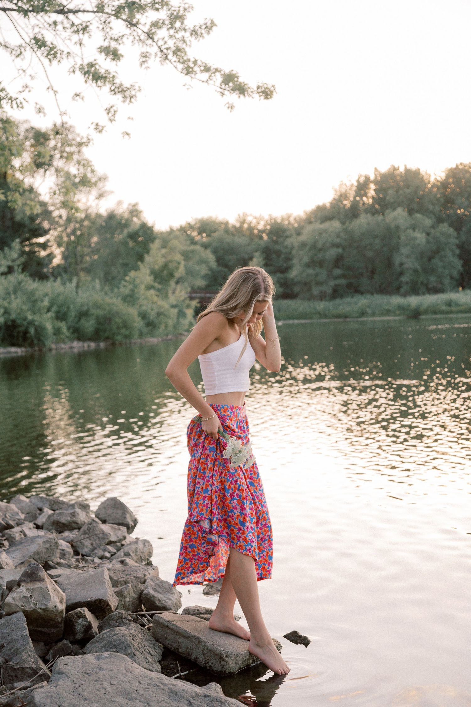 Person wearing a floral skirt walks along rocky lakeside during sunset.