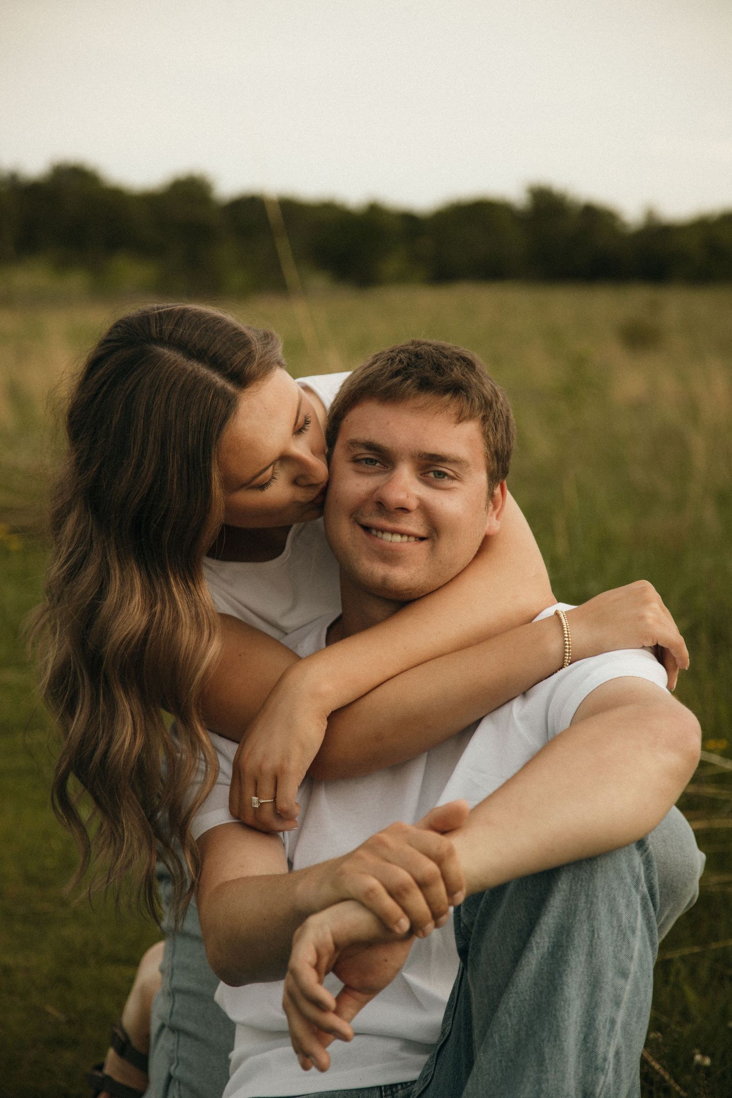 A smiling man gives a piggyback ride to a woman hugging him from behind in a green field.