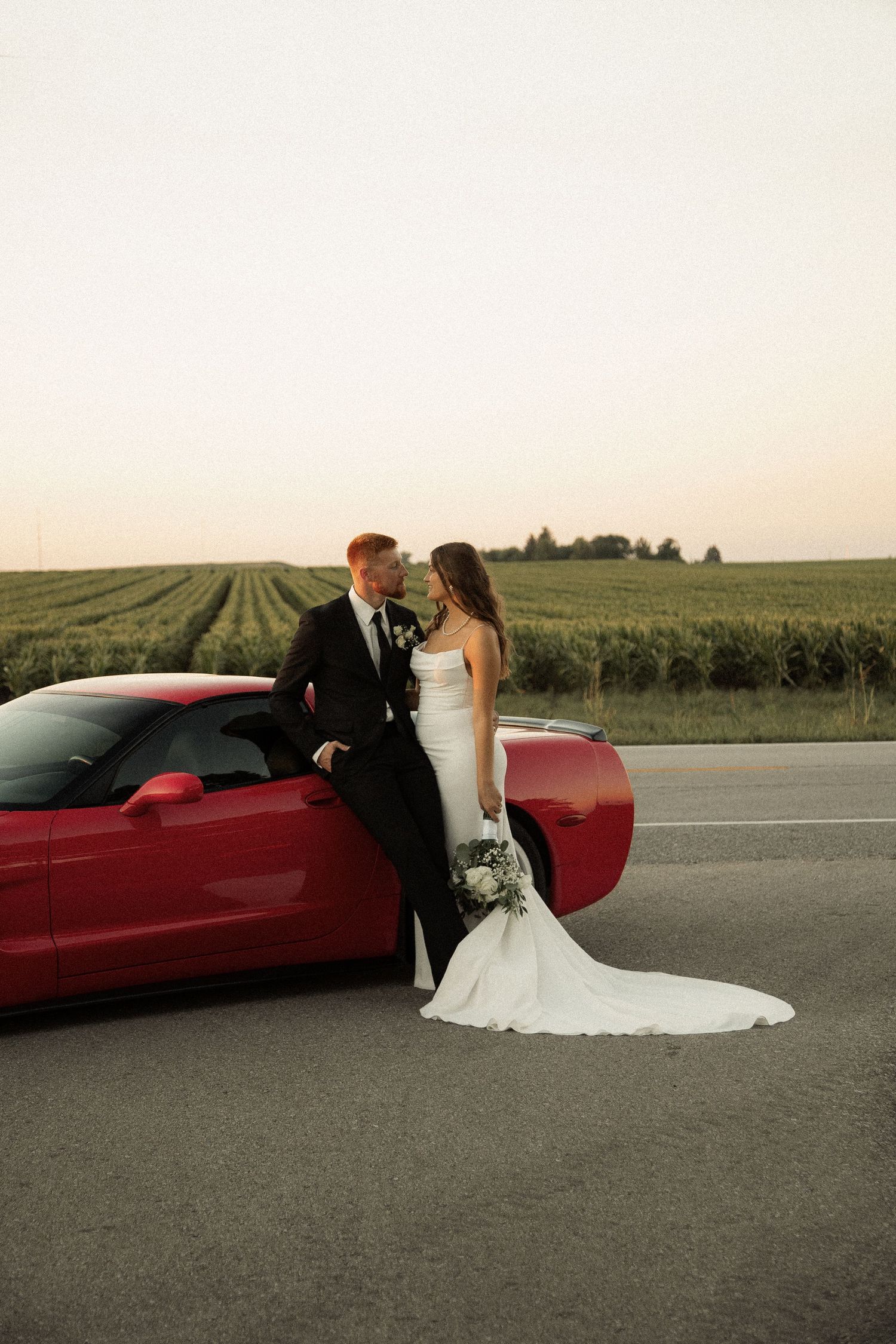 Newlyweds pose beside a red sports car with rolling fields in the background at sunset.