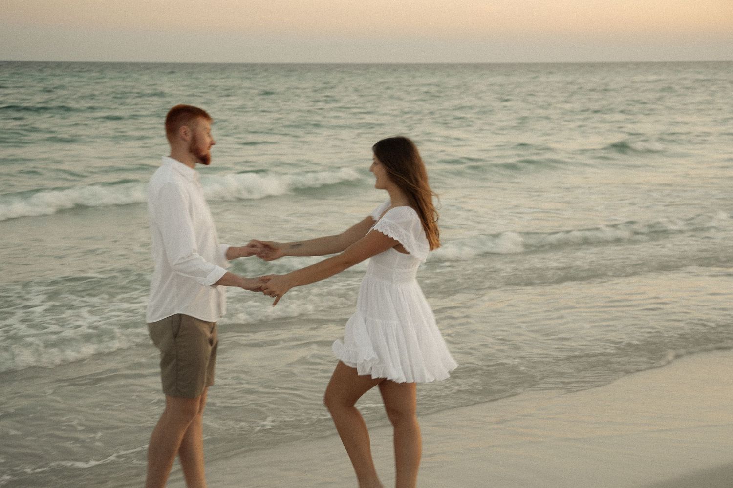 Couple in white clothing dancing playfully on beach at dusk.