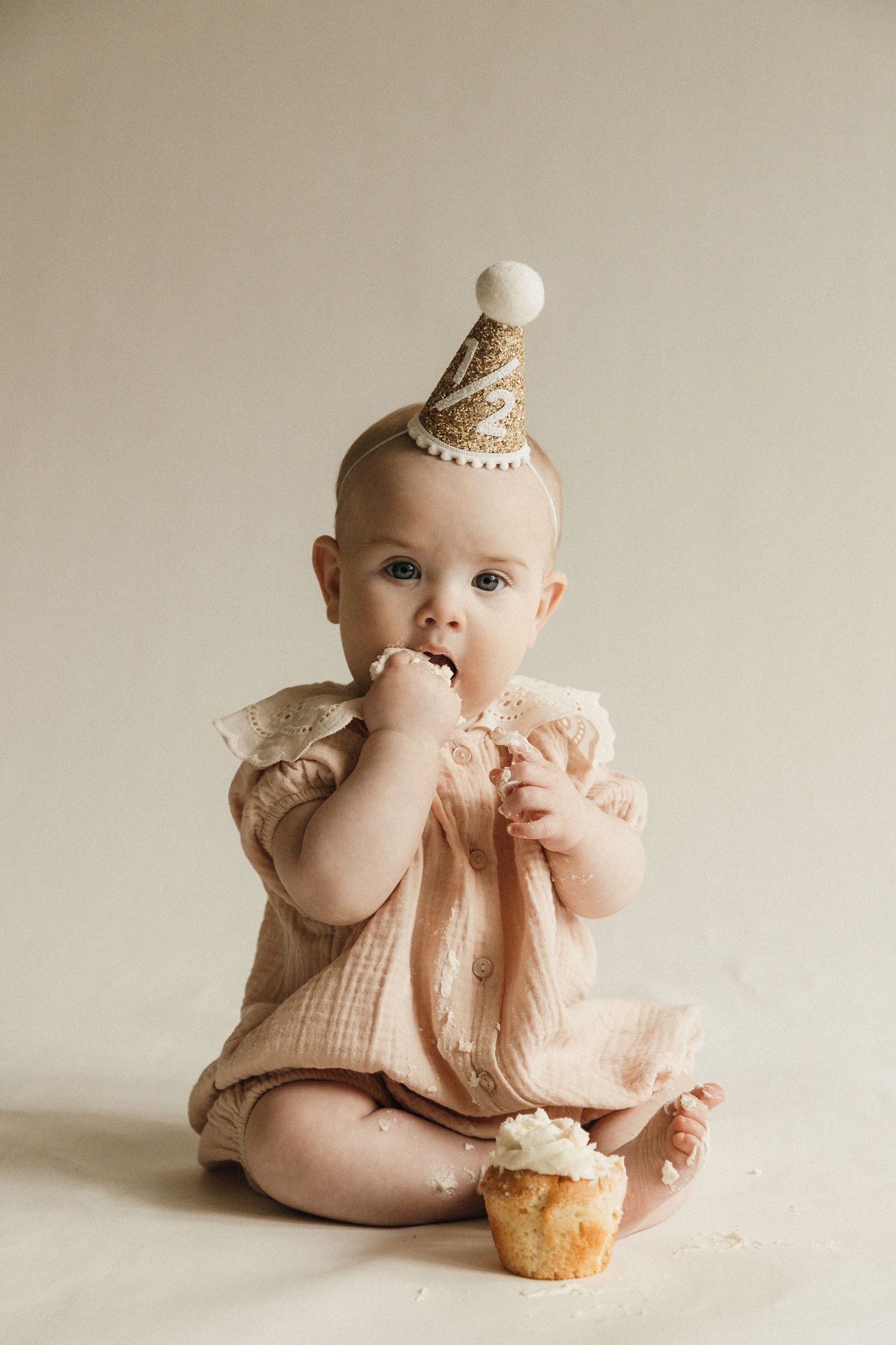 Adorable baby girl in pink romper and party hat eating her first birthday cupcake.