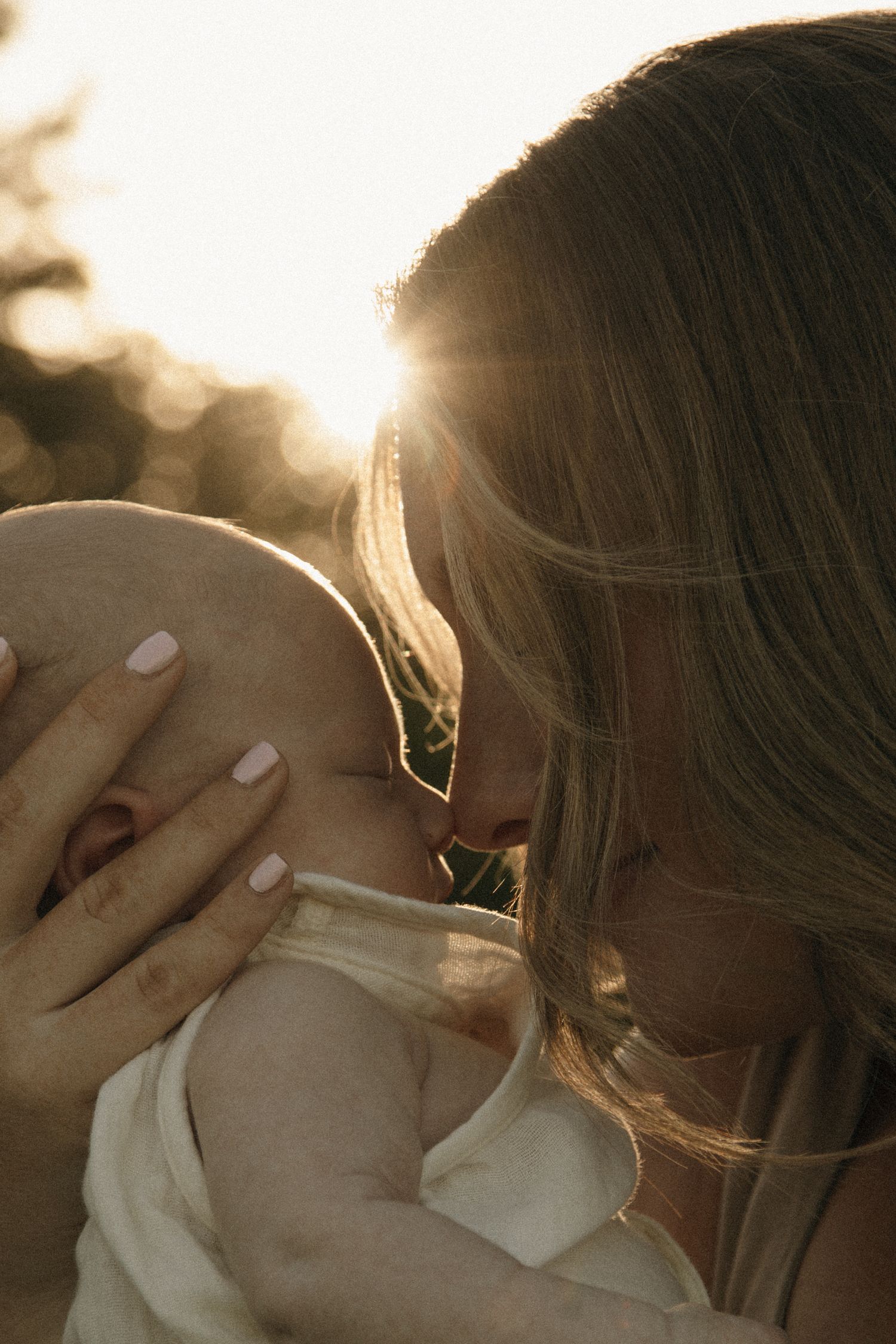 A mother tenderly kisses her newborn baby in golden backlit sunlight outdoors.
