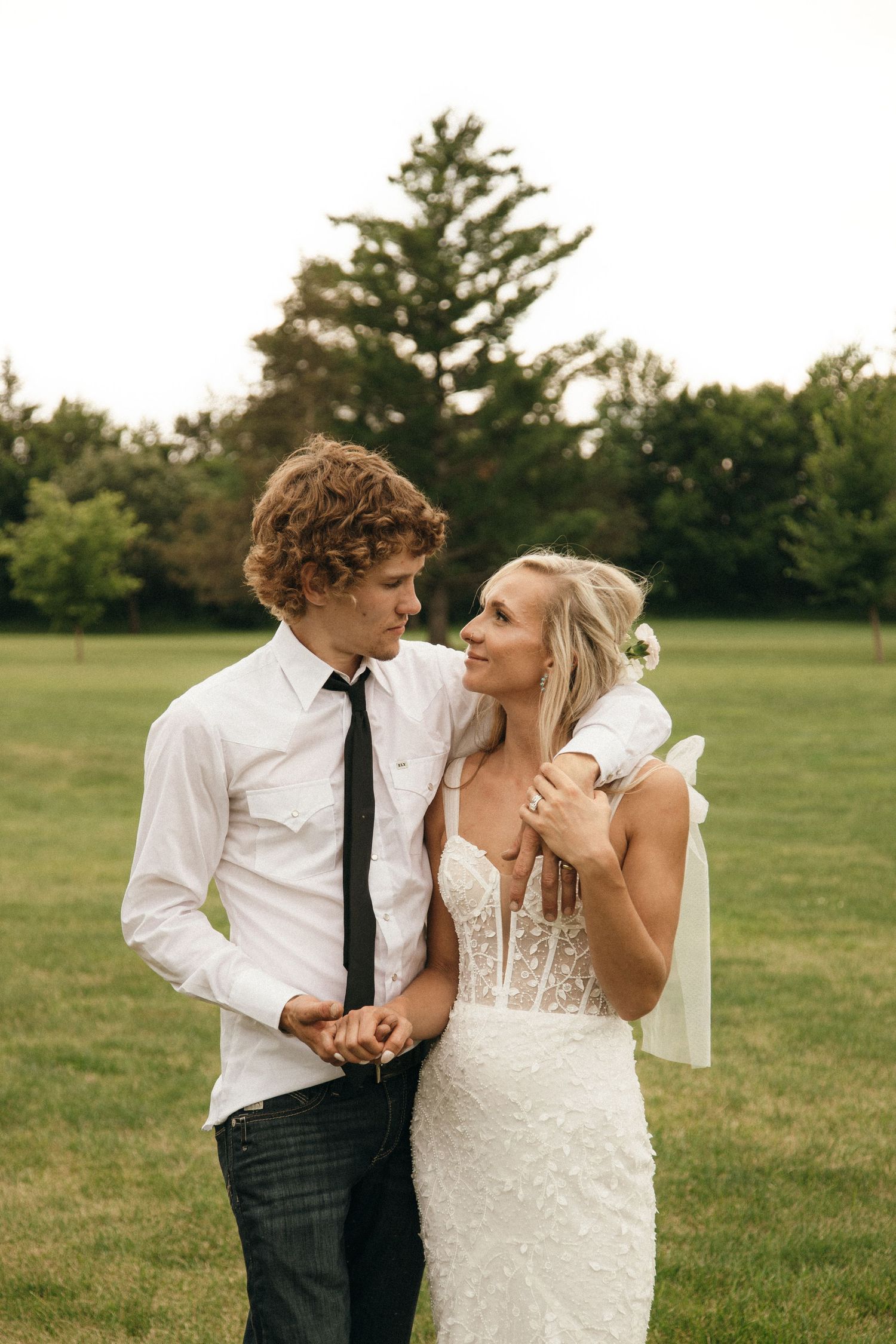 Bride in a beaded gown and groom in white shirt and black tie share a tender moment on a green lawn.