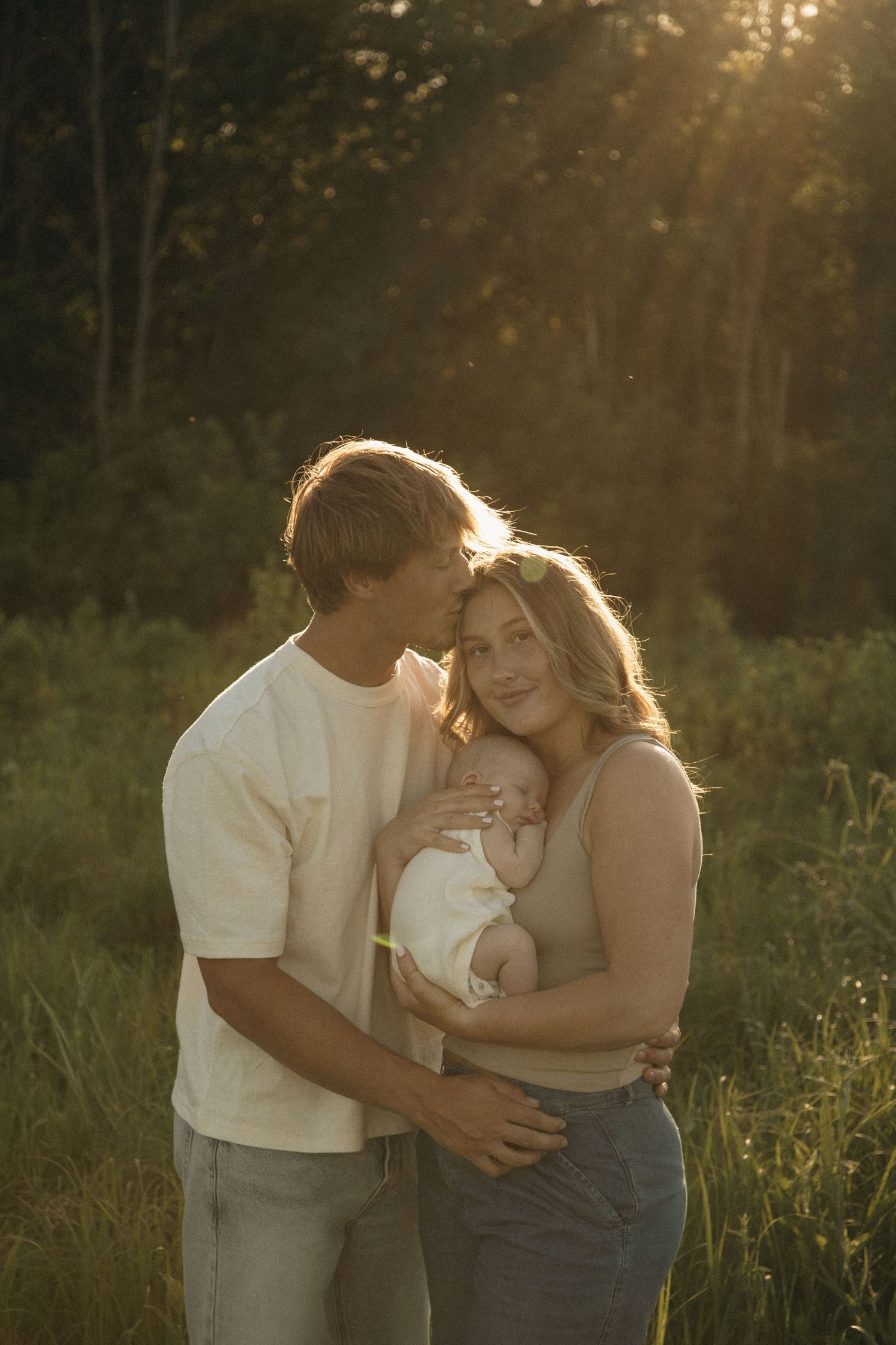 Young couple holding newborn baby outdoors in golden sunset light, standing in green meadow with trees in background.