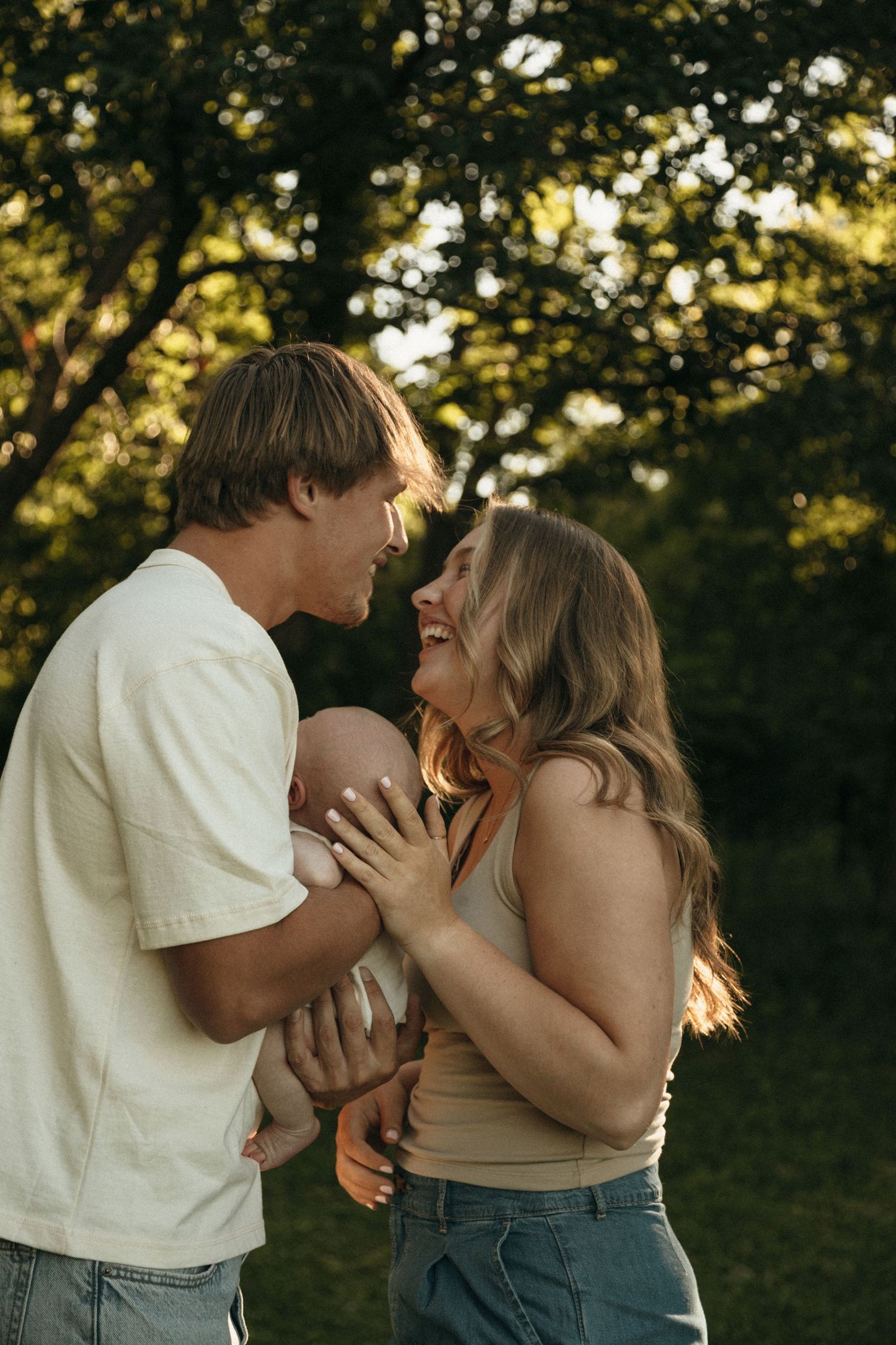 Happy couple laughing together while holding their newborn baby in a sunlit park.