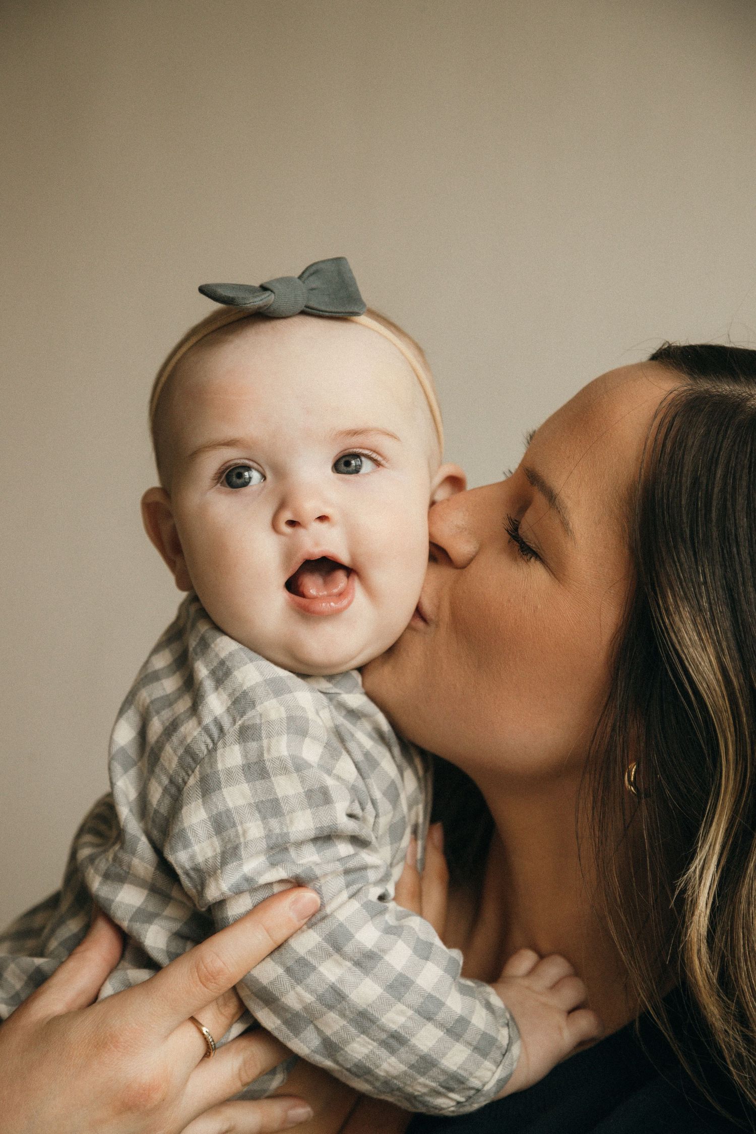 A mother tenderly kisses her smiling baby girl wearing a gray bow headband and plaid outfit.