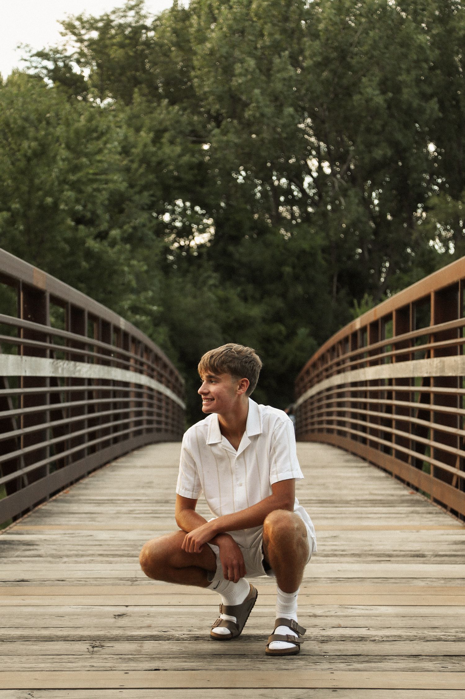 Person in white shirt relaxing on a wooden footbridge with trees in background.