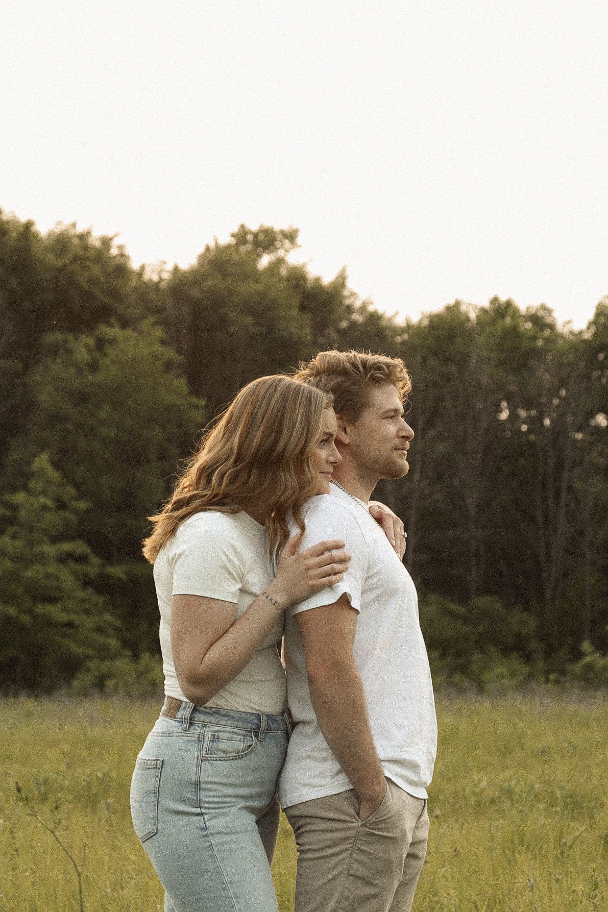 A couple in casual attire standing close together in a sunlit meadow with trees in the background.