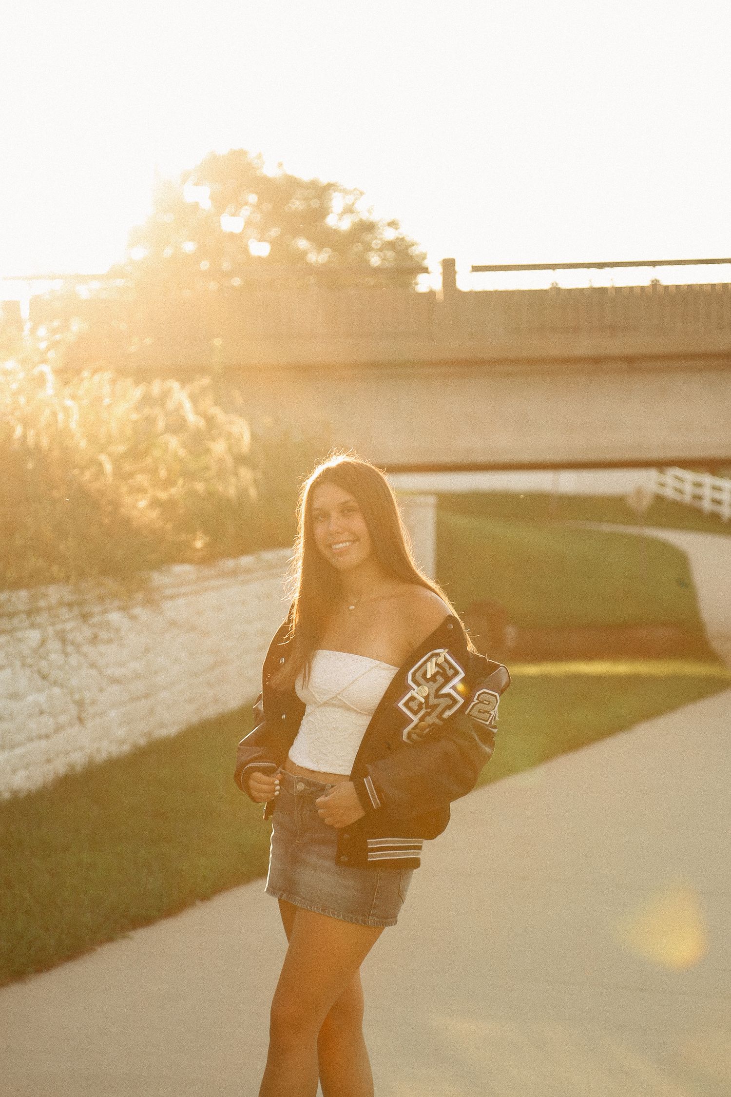 A person in a varsity jacket poses against a sunset-lit backdrop with golden light filtering through.