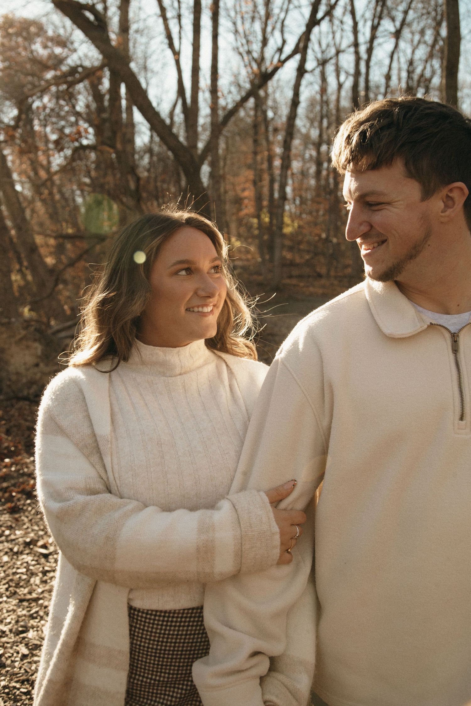 A romantic autumn photoshoot showing a couple in white sweaters laughing together in a wooded setting.