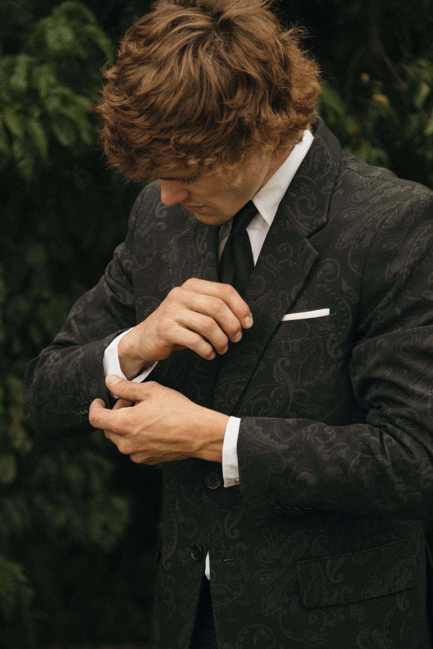 Elegant man in patterned dark suit adjusting cufflinks outdoors, wearing black tie and white pocket square.