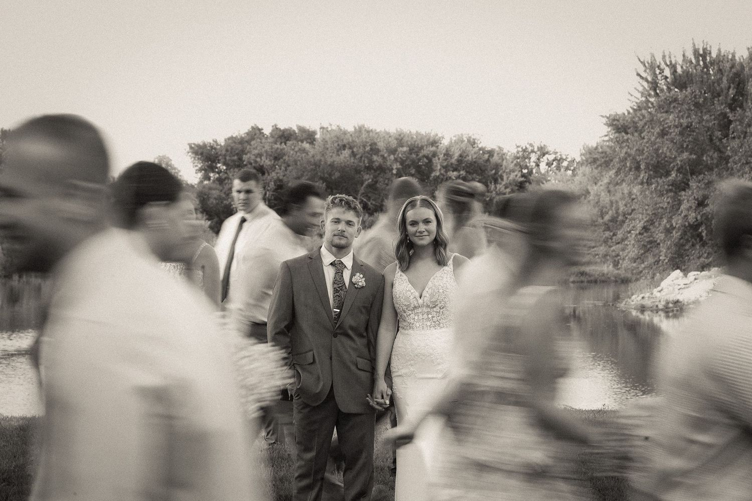 A wedding scene with motion-blurred guests surrounding the couple by a lake.
