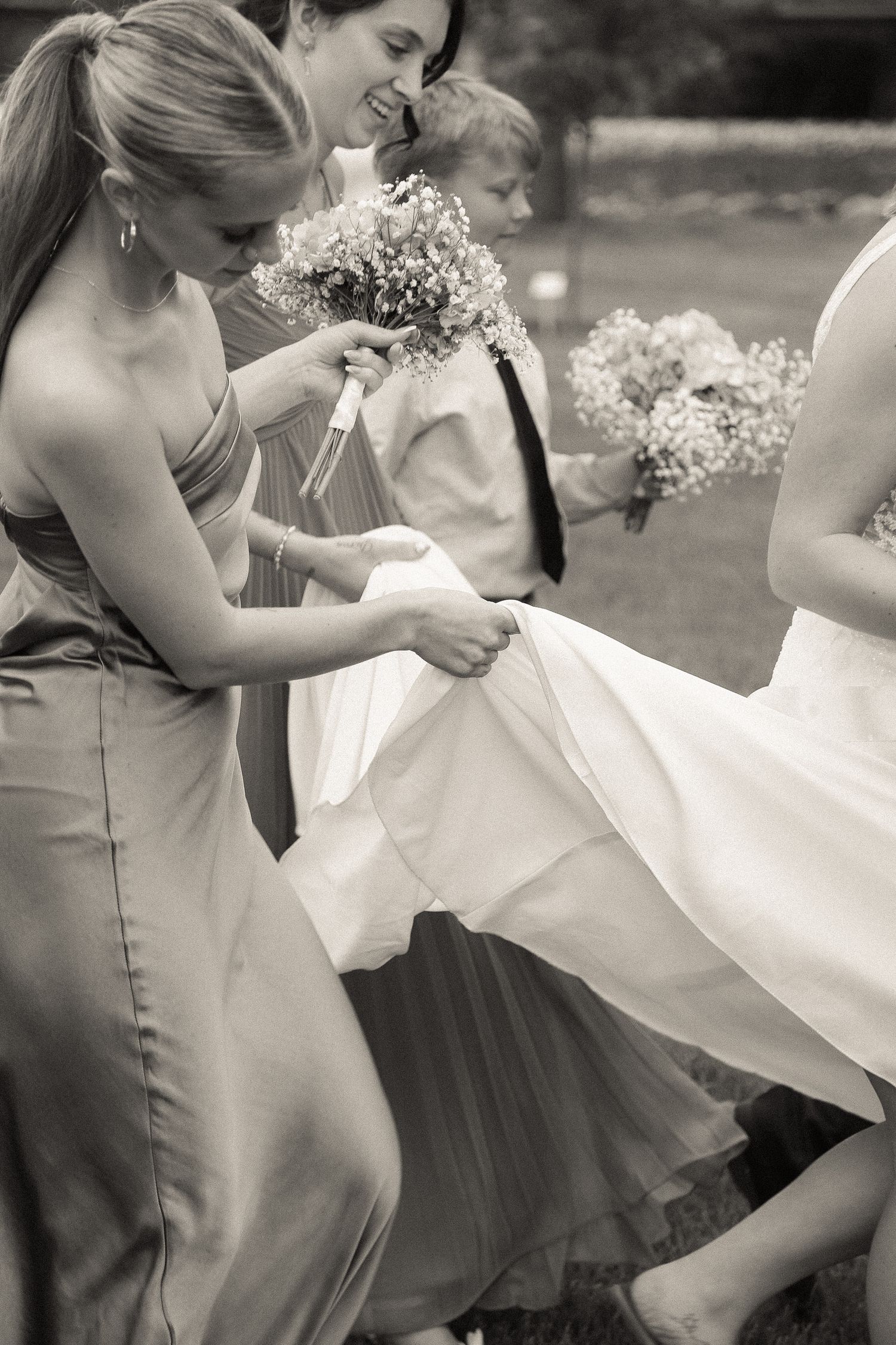 Bridesmaids in elegant dresses help adjust the bride's gown outdoors.