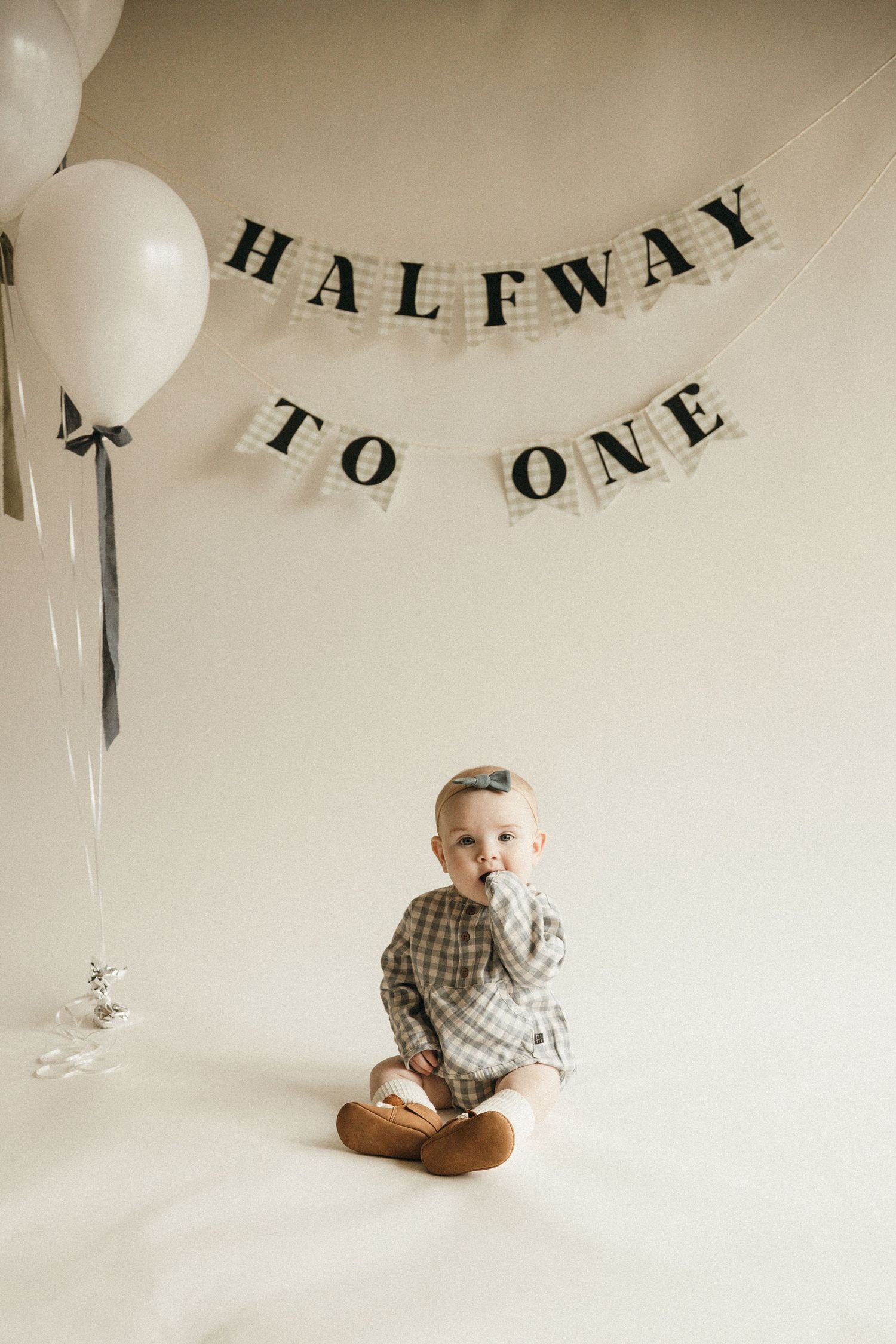 Baby in plaid romper sits before a 'Halfway to One' banner with white balloons, celebrating a 6-month milestone.