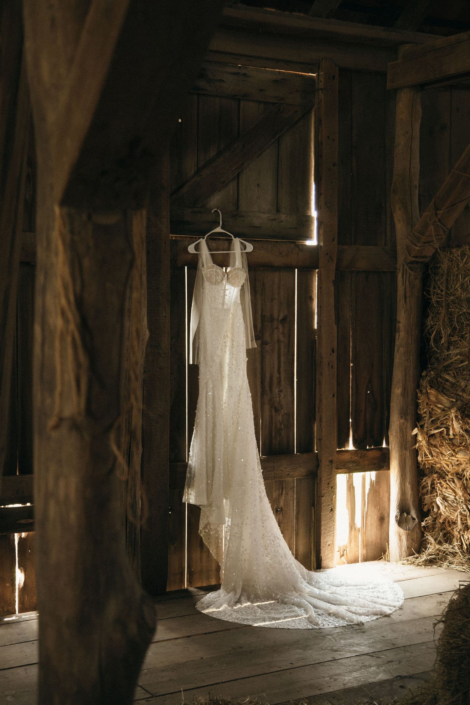 White lace wedding dress hanging in rustic barn with wooden beams and hay bales in warm light.