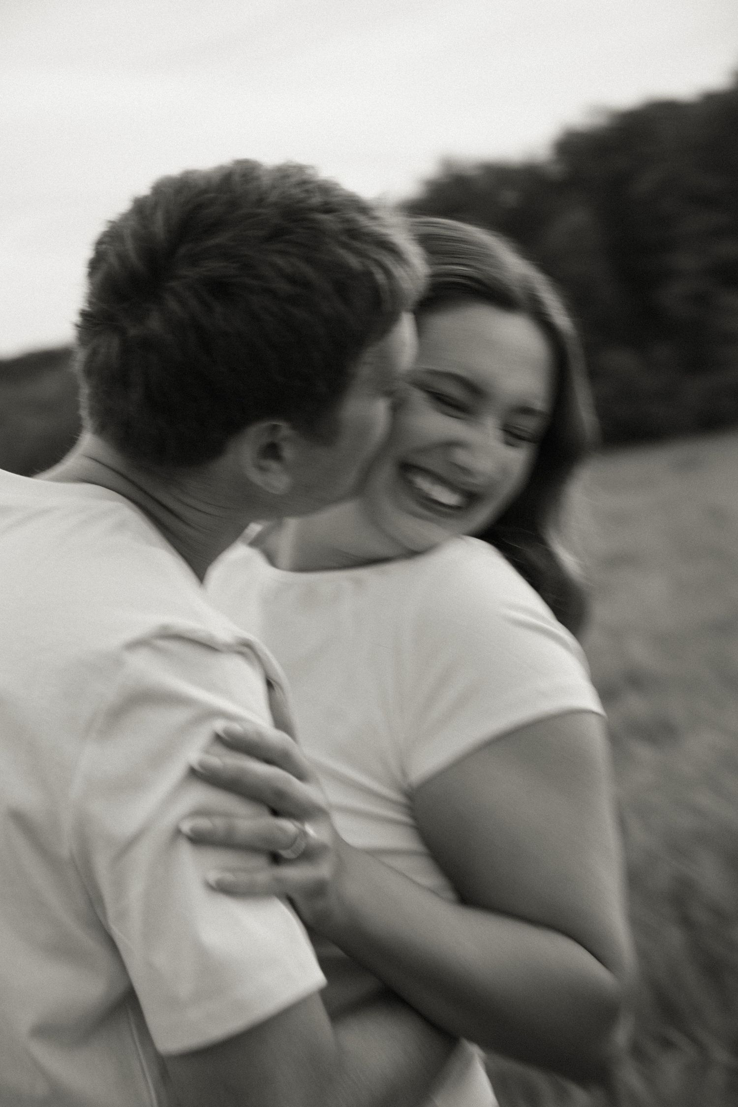 Black and white image of a laughing woman being kissed on the cheek by her partner outdoors.