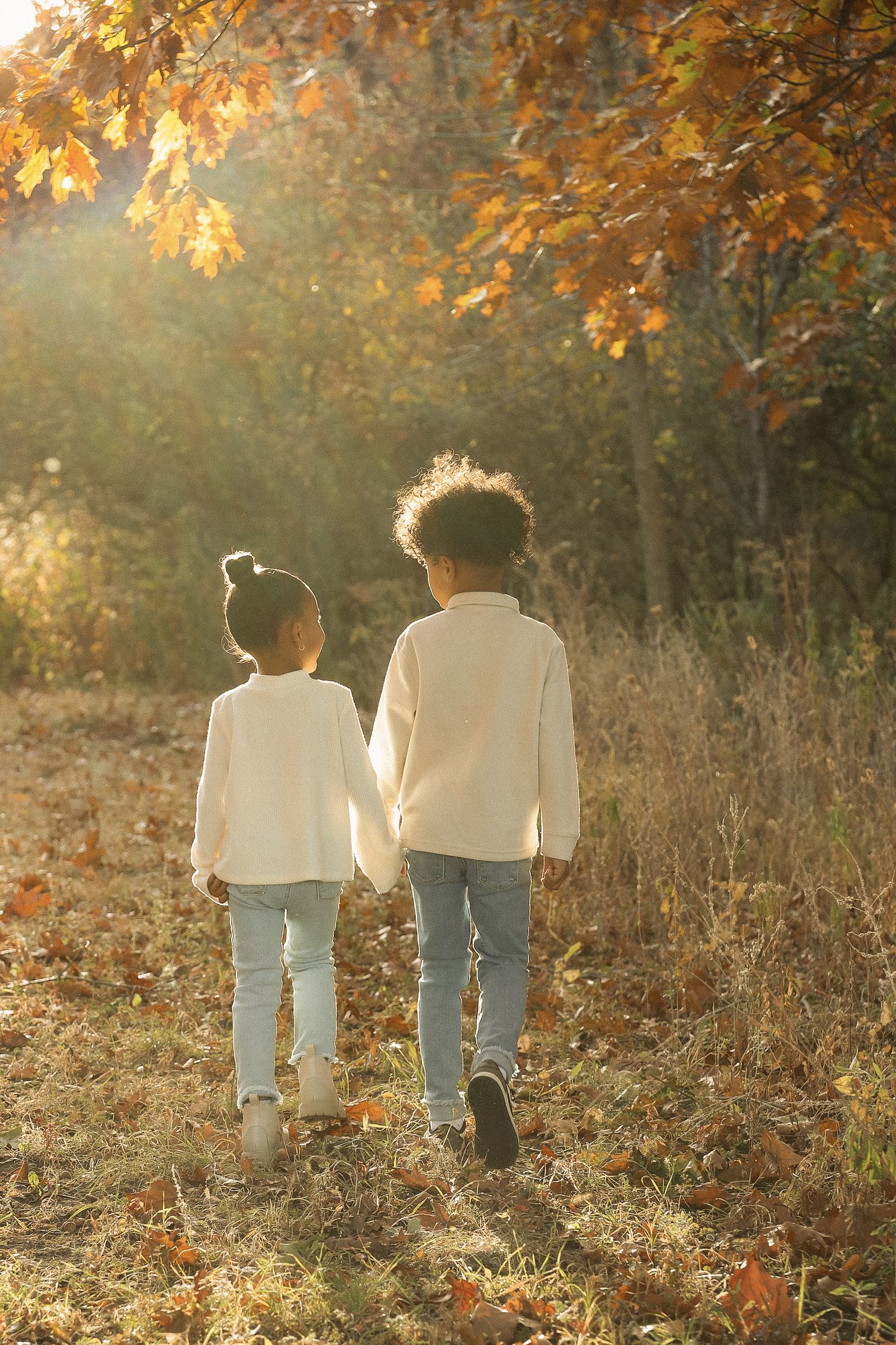 Two children in matching white sweaters walk through a sunlit autumn forest.