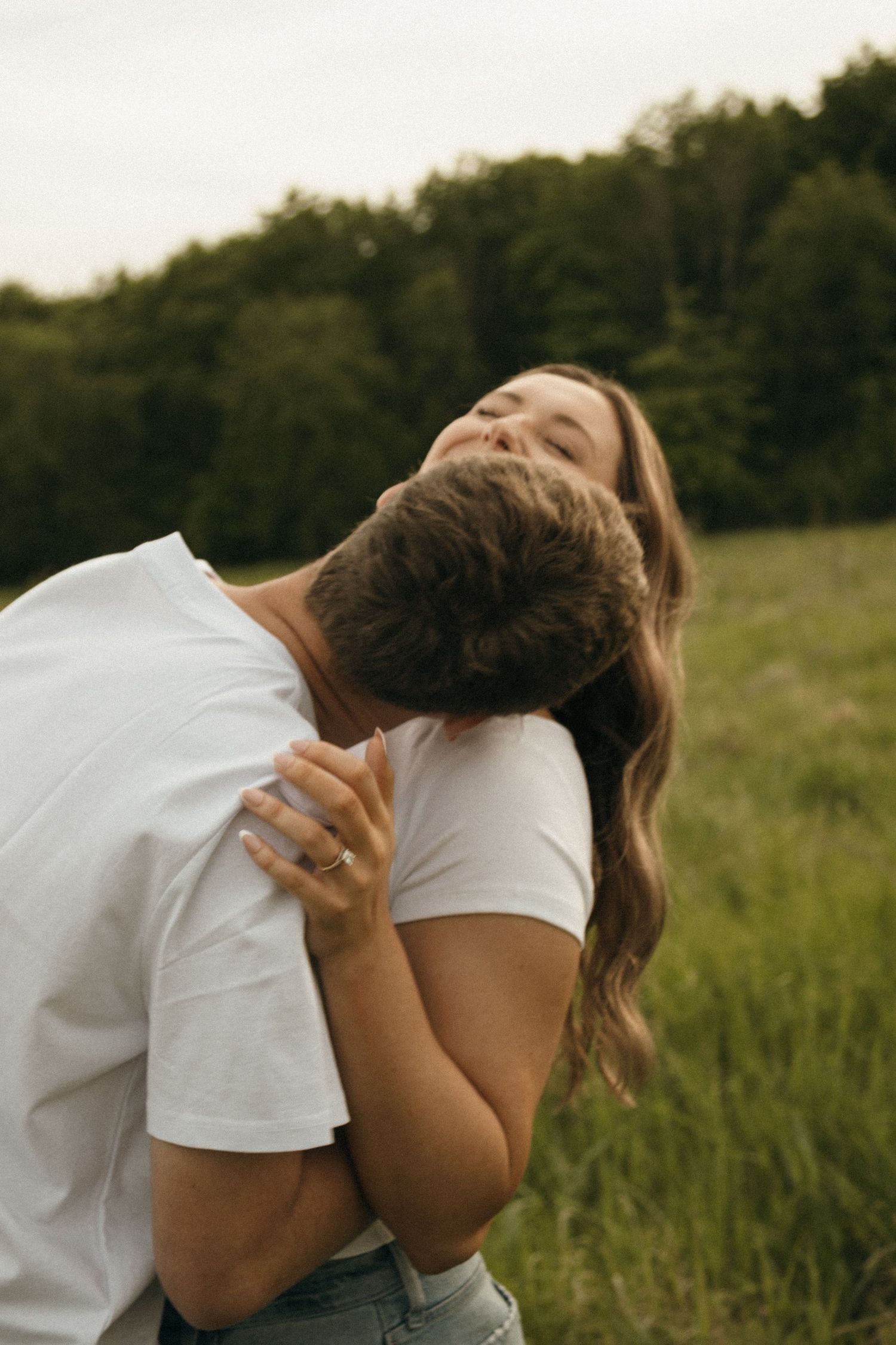 A joyful woman hugs a man in a white t-shirt while laughing in a sunny green meadow.