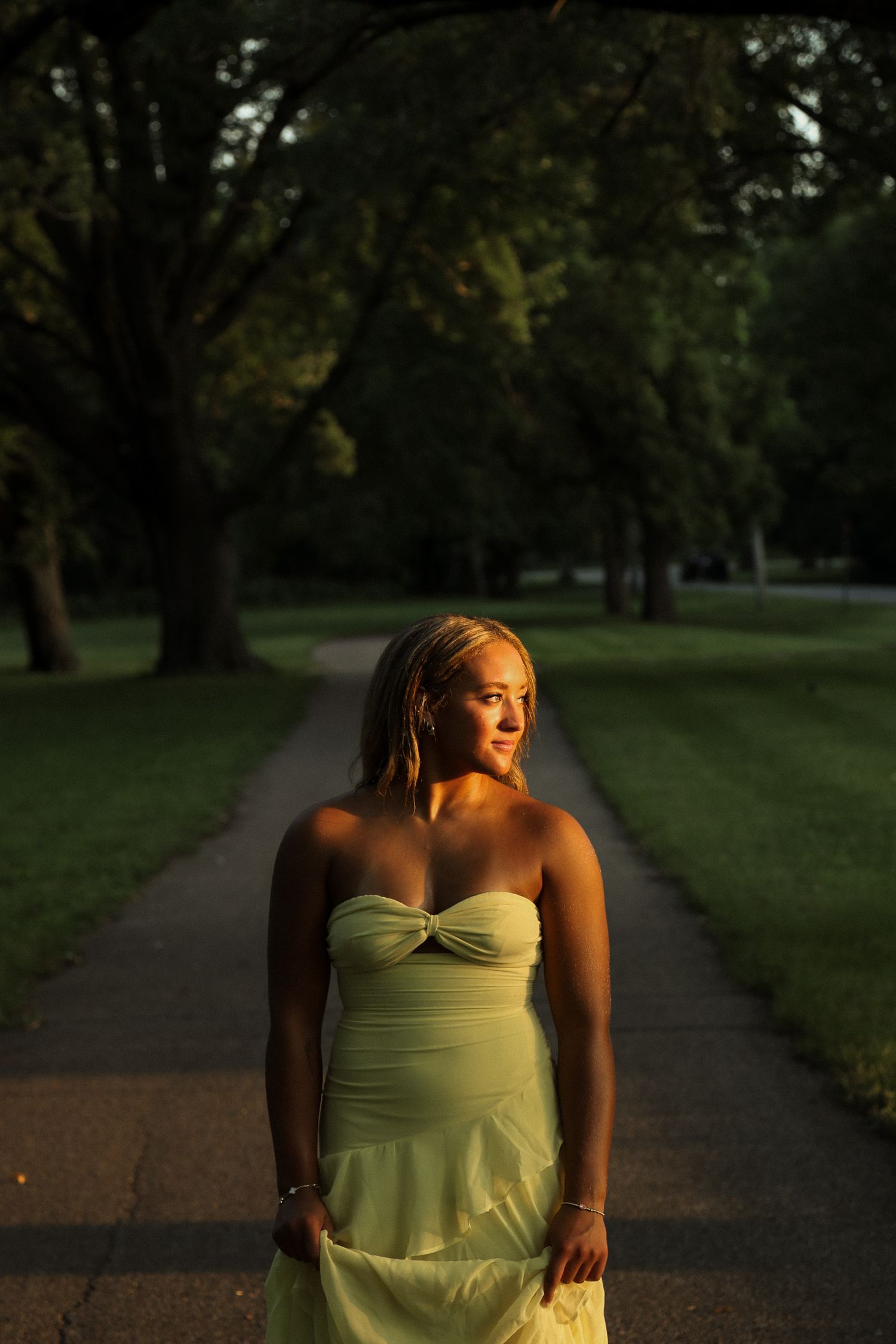Dramatic sunset lighting illuminates someone in a yellow dress on a tree-lined path.