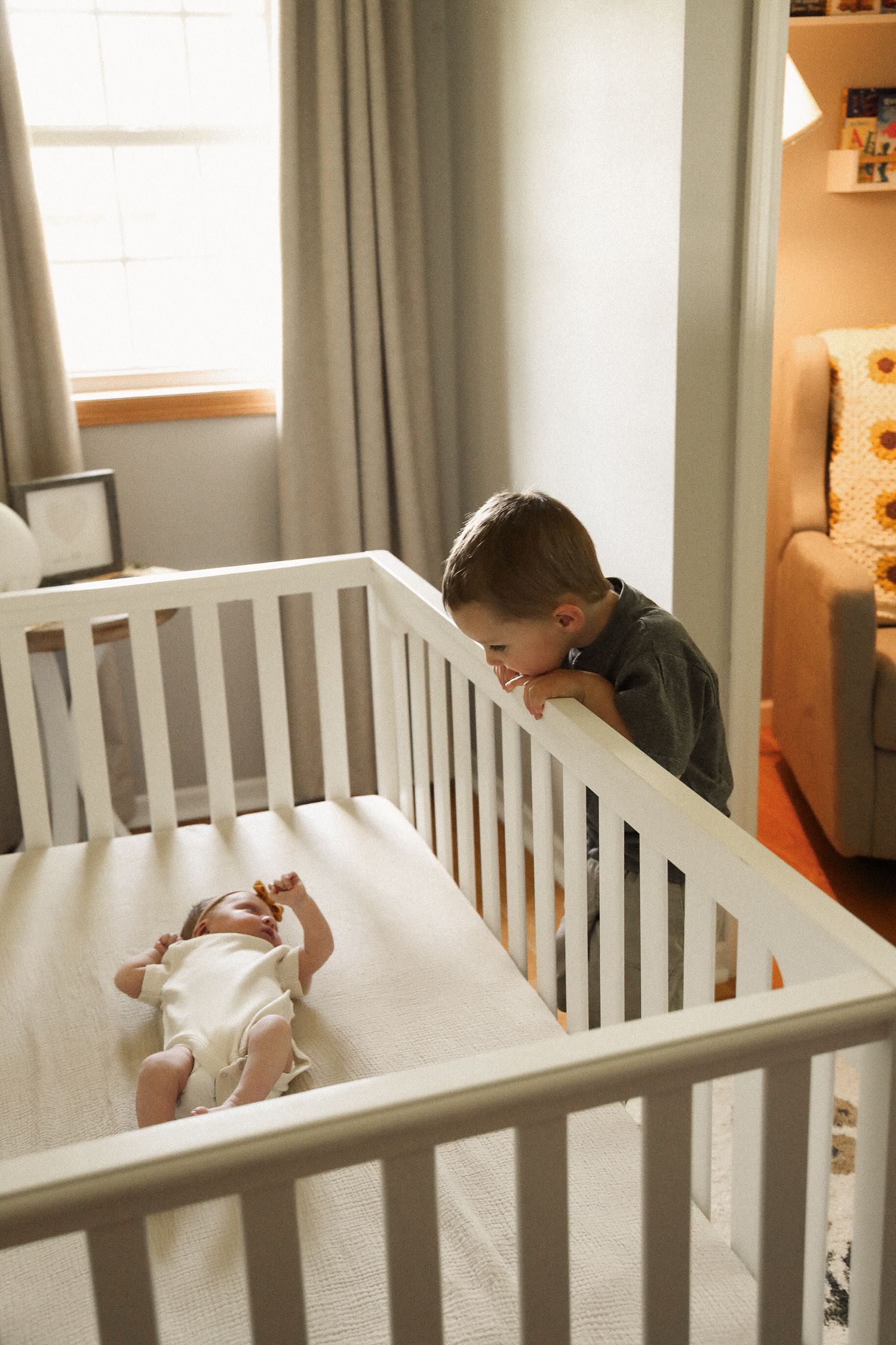Parents stand over a white crib watching their sleeping baby in a sunlit room.