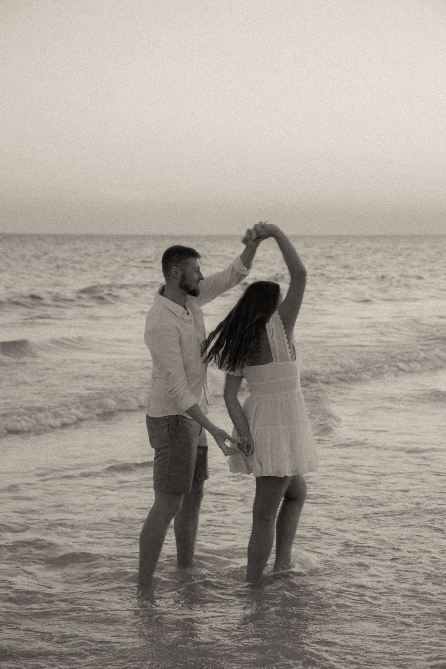 A couple dances in shallow ocean water, creating a romantic black and white silhouette against the waves.