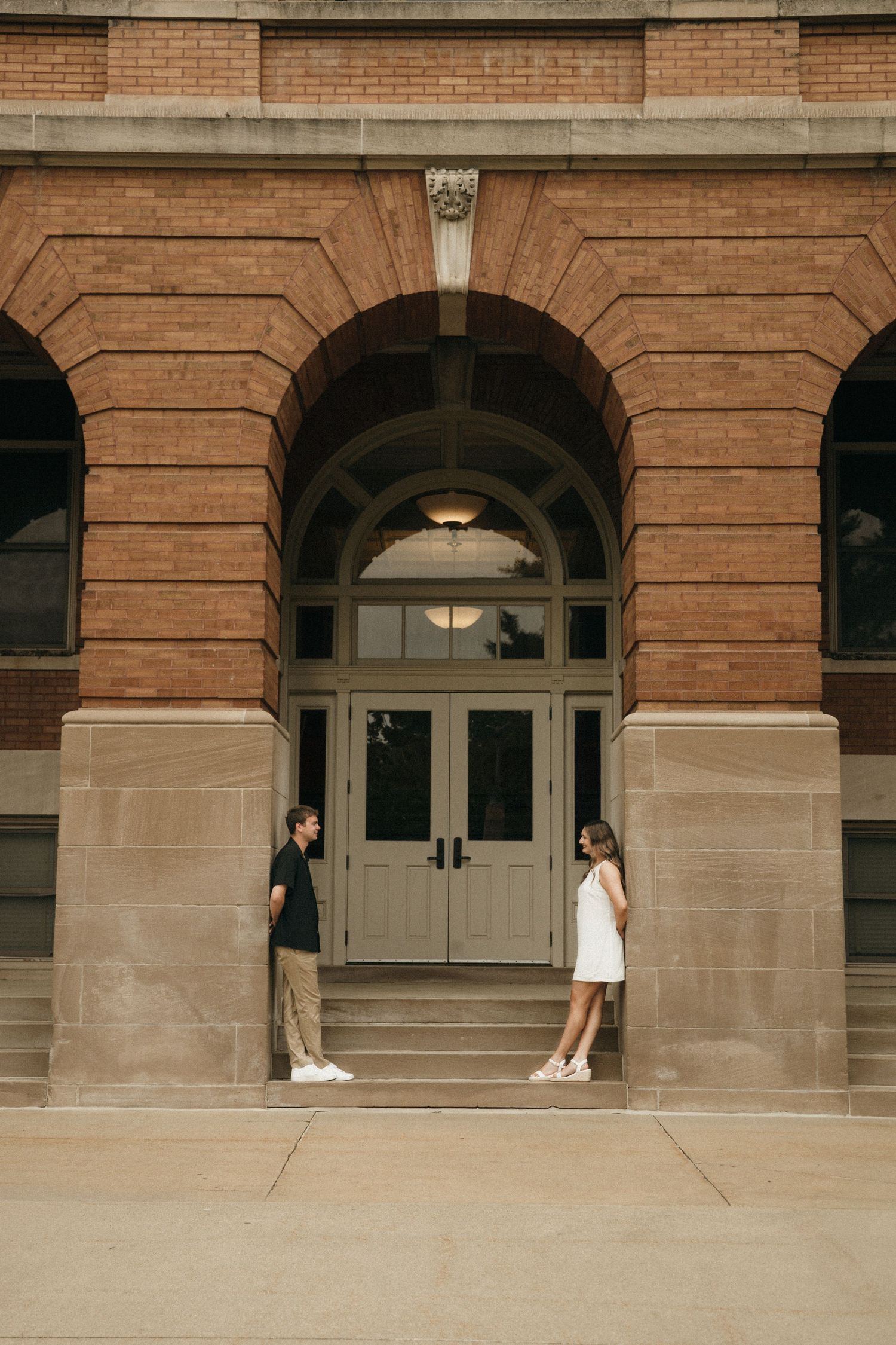 A couple stands facing each other at the entrance of a historic brick building with an arched doorway.