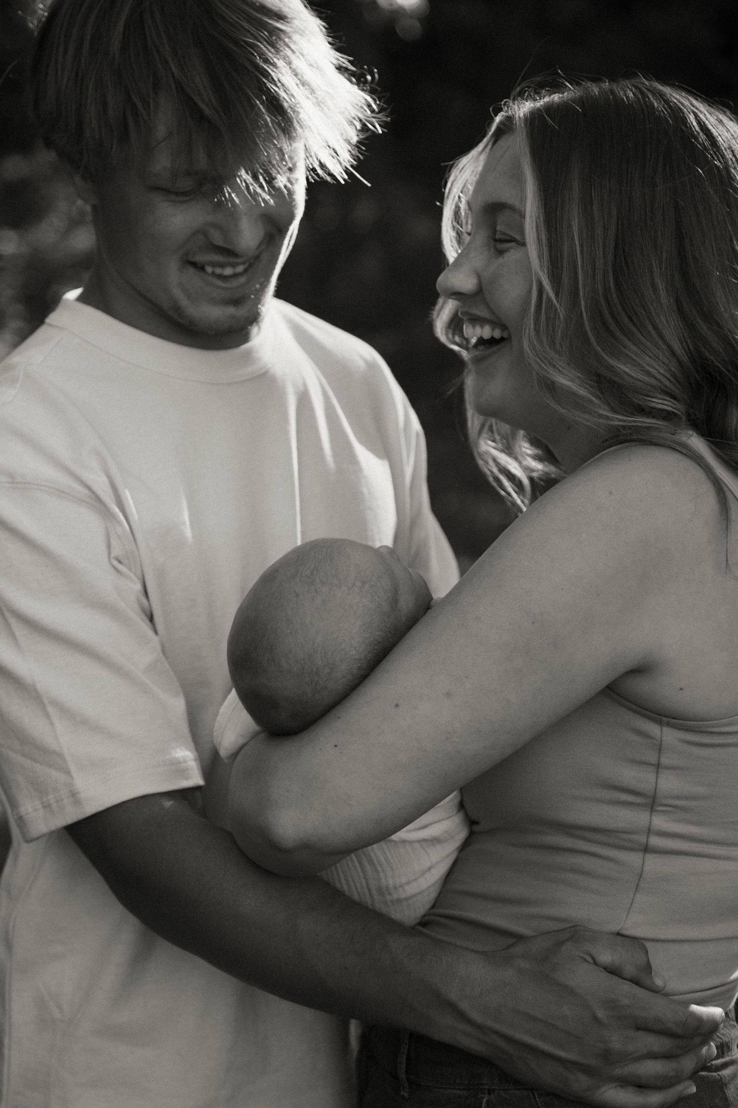 A laughing couple shares a joyful moment together holding their newborn baby in a black and white outdoor photo.