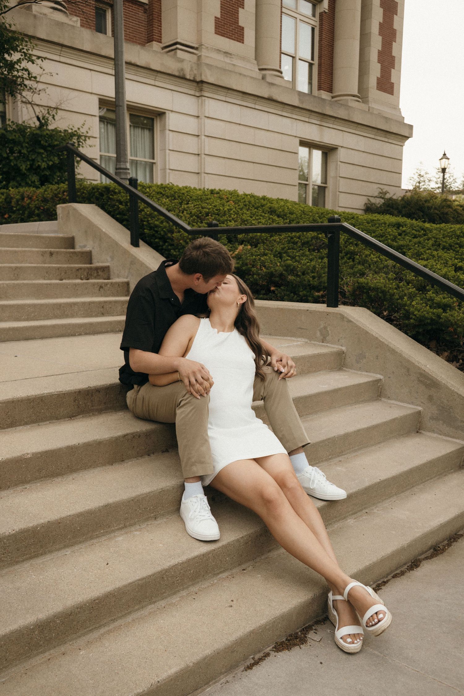 Couple sharing a romantic kiss while sitting on outdoor stone steps near a classic brick building.