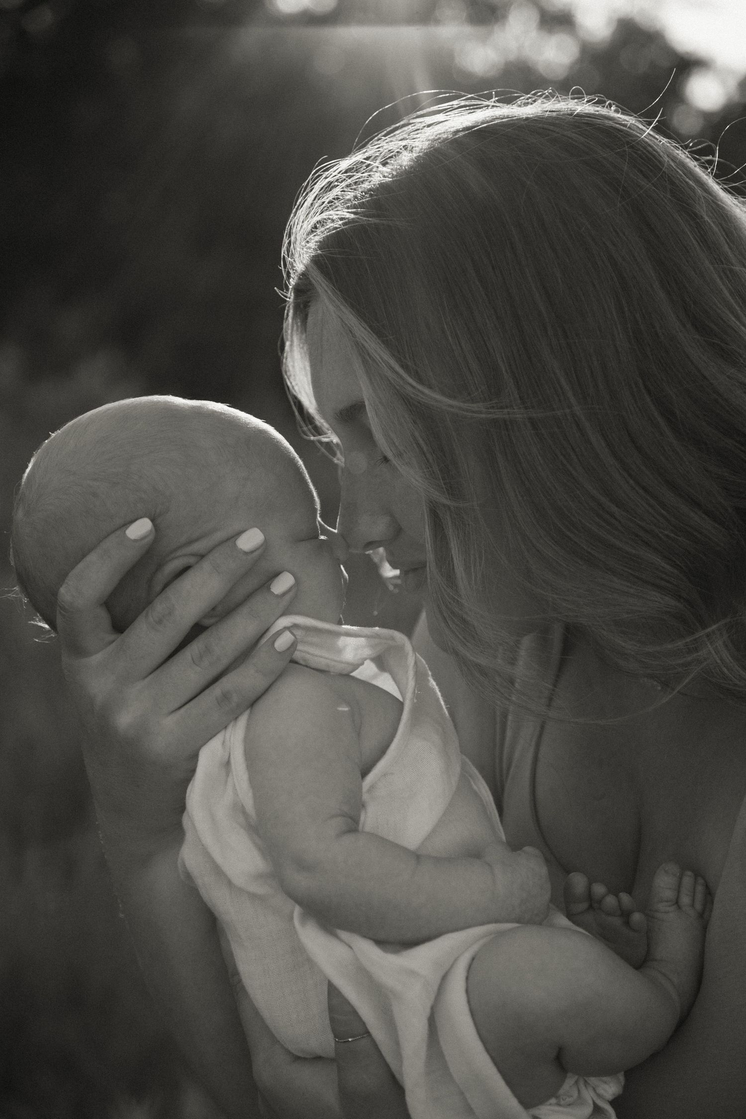 Black and white photo of mother gently kissing her newborn baby outdoors in soft sunlight.