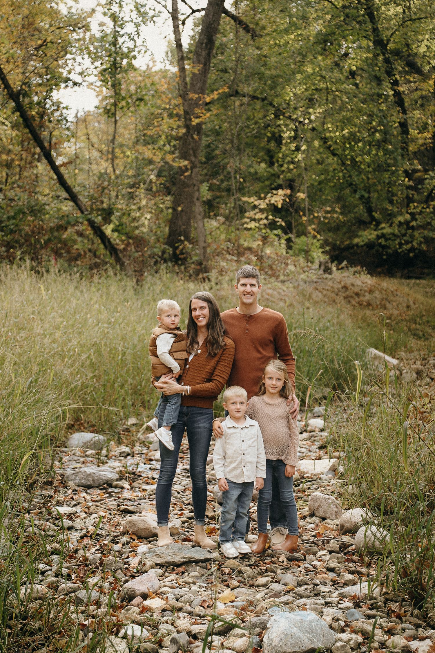 Family walking together on rocky path through autumn trees in earth-toned clothing.