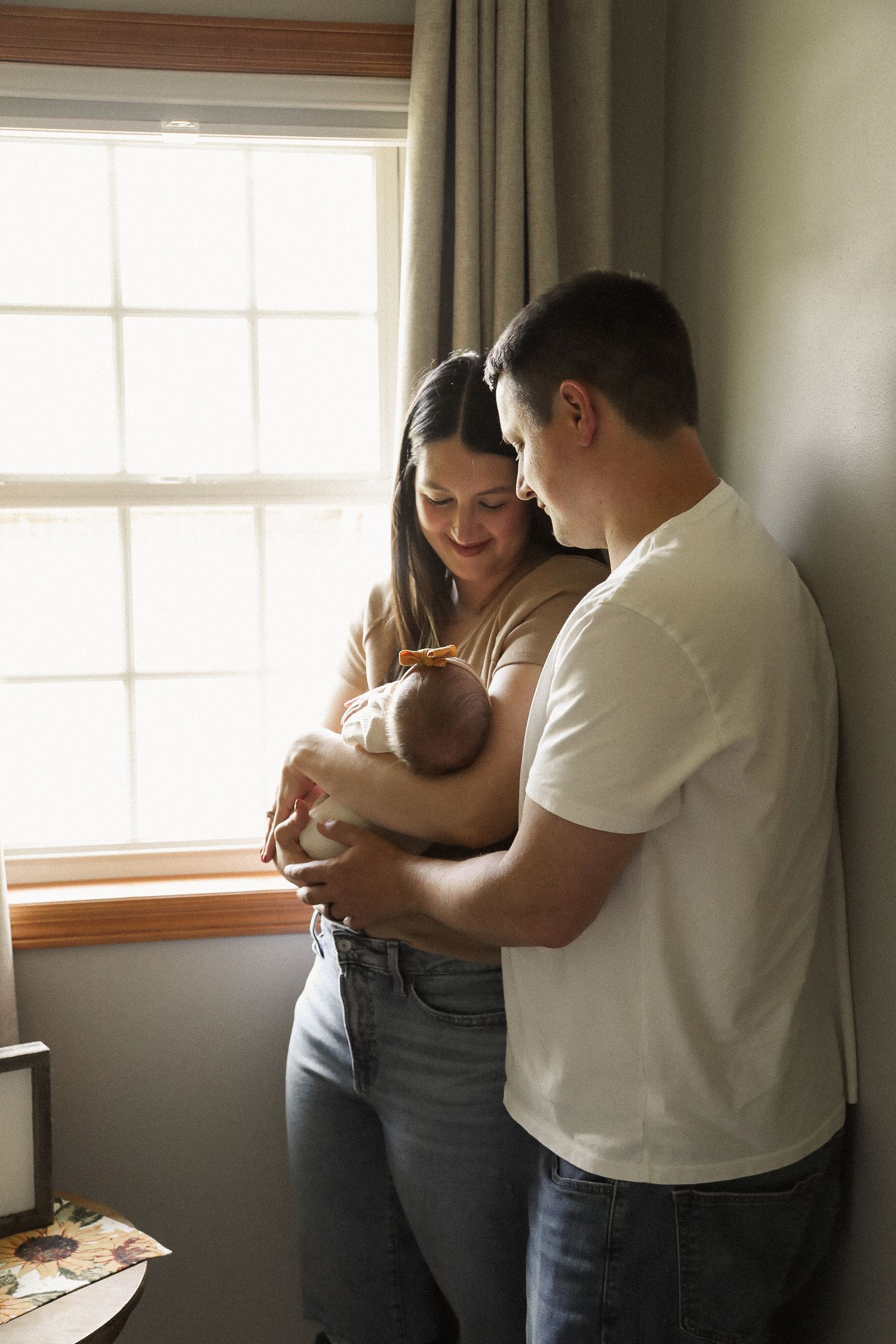 A couple embraces their baby in a sunlit room with grey walls and window.