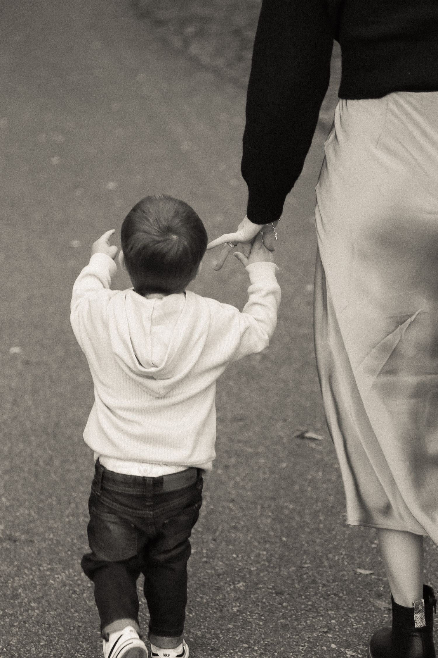 A small child in a white shirt and dark pants holds onto an adult's leg while walking.