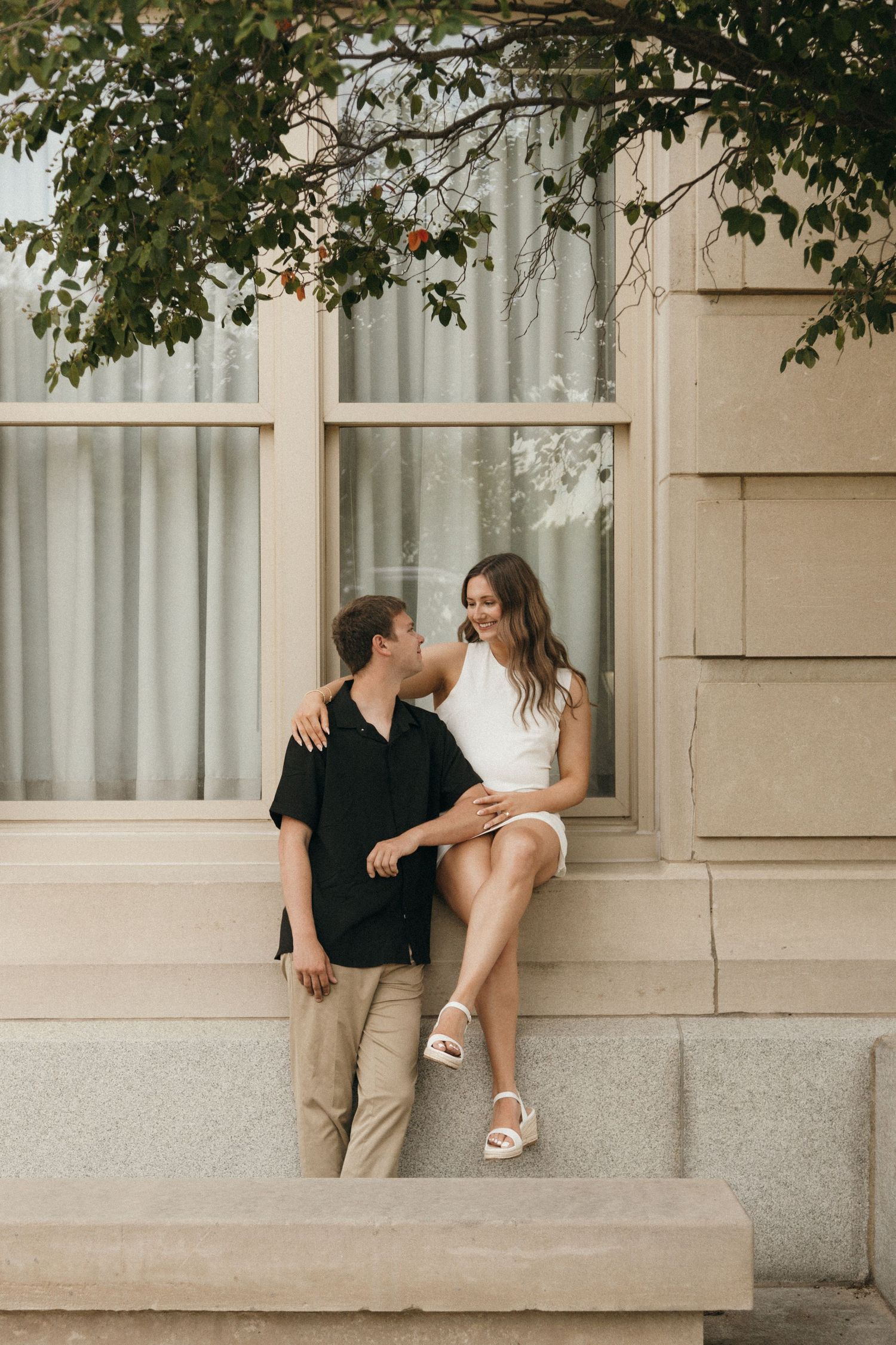 Happy couple sitting on stone steps outside building, smiling at each other under tree.