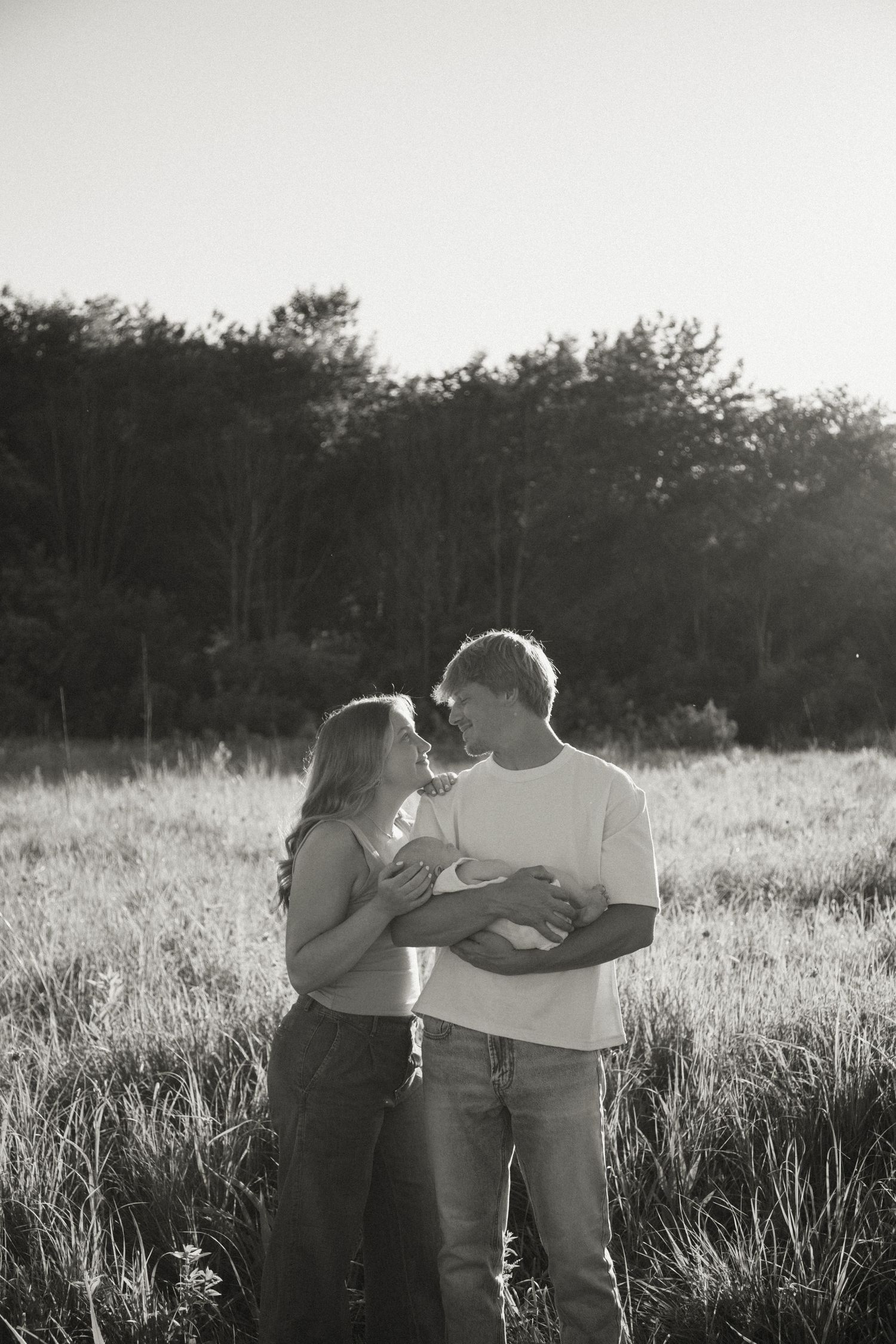 Black and white photo of a couple holding a newborn baby in a grassy field with trees in the background.
