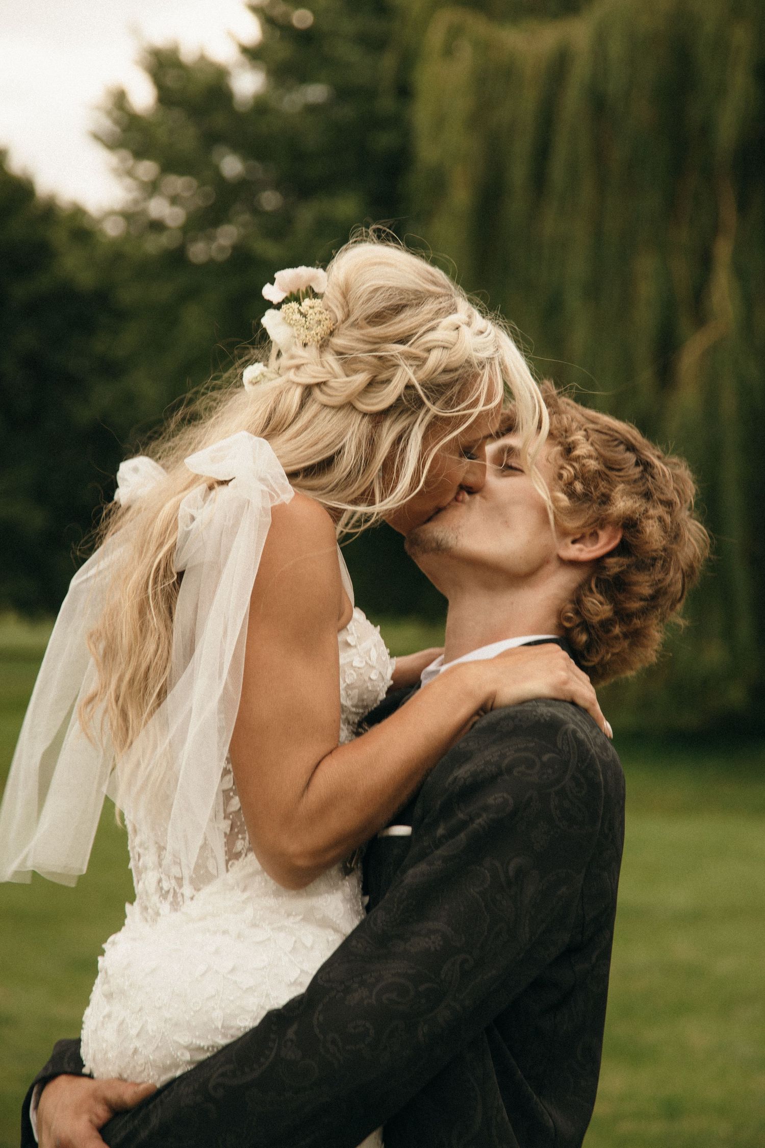 Bride and groom share a romantic kiss outdoors, with the groom lifting the bride in her lace wedding dress and floral veil.