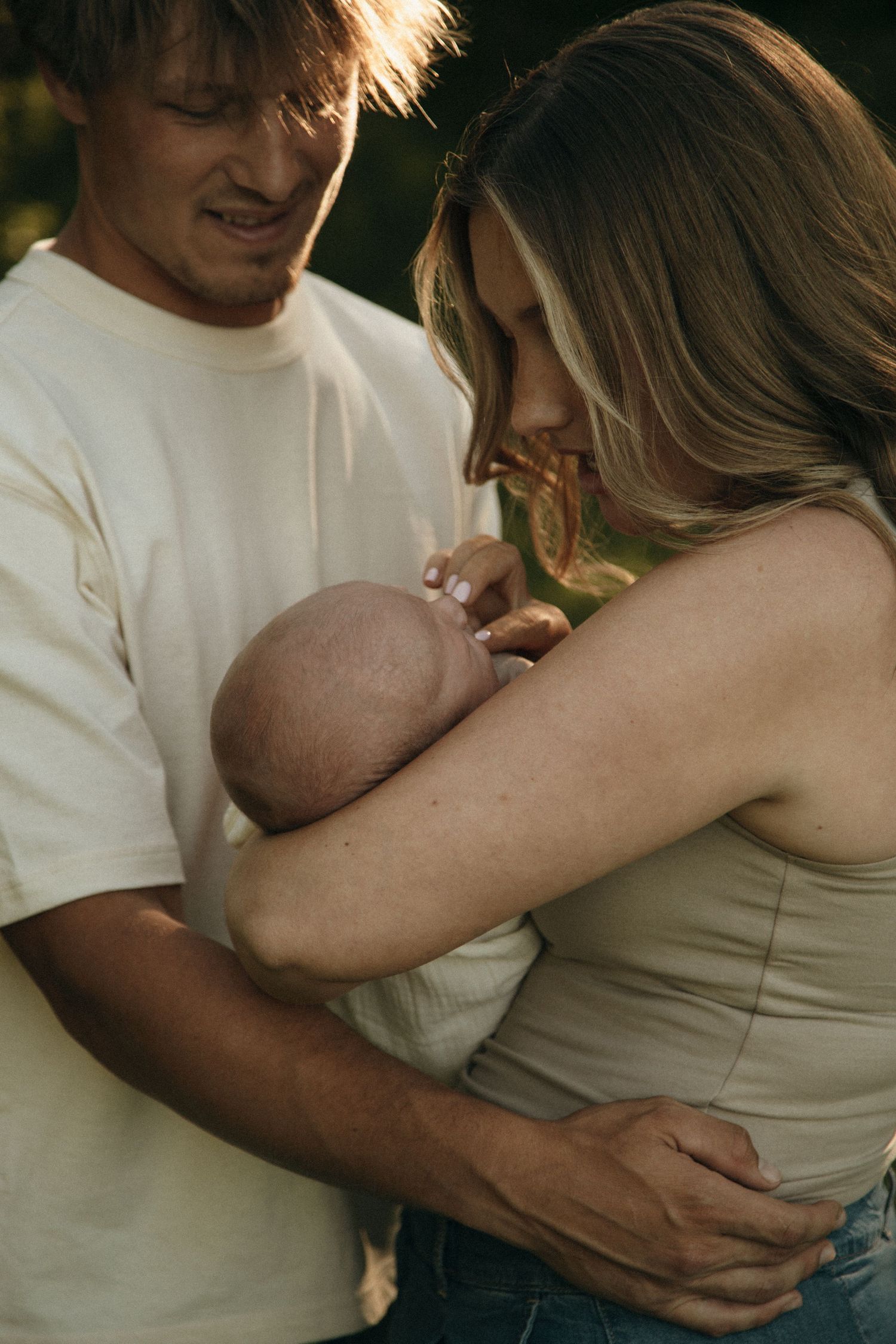 Young parents lovingly cradle their newborn baby together outdoors in warm golden light.