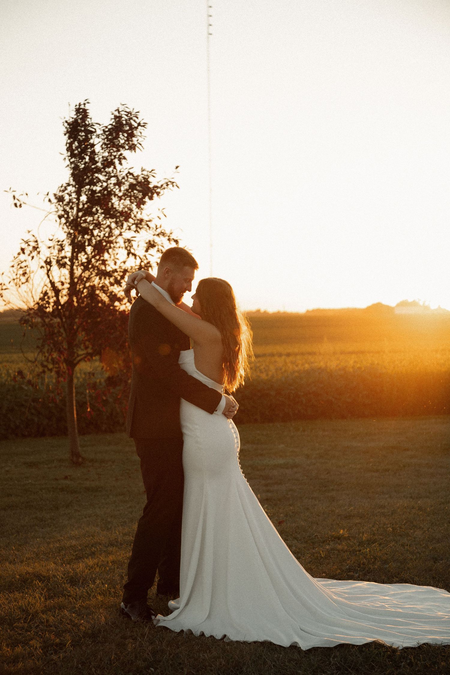 Silhouetted newlyweds embrace in front of a setting sun with a flowing white wedding dress train.