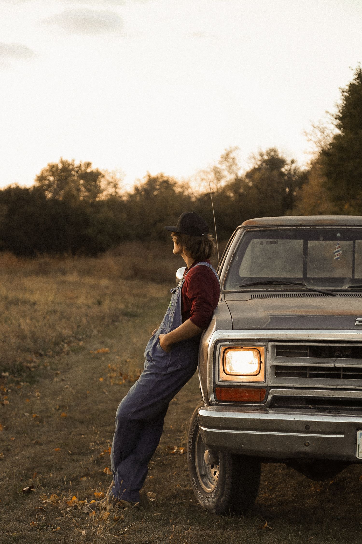 Person in overalls poses near headlights of vintage Dodge truck at dusk.
