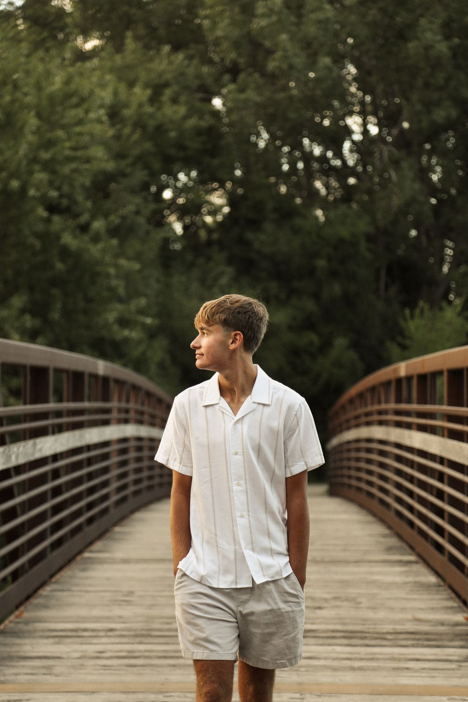 A person in white clothing sits casually on a wooden bridge surrounded by lush greenery.